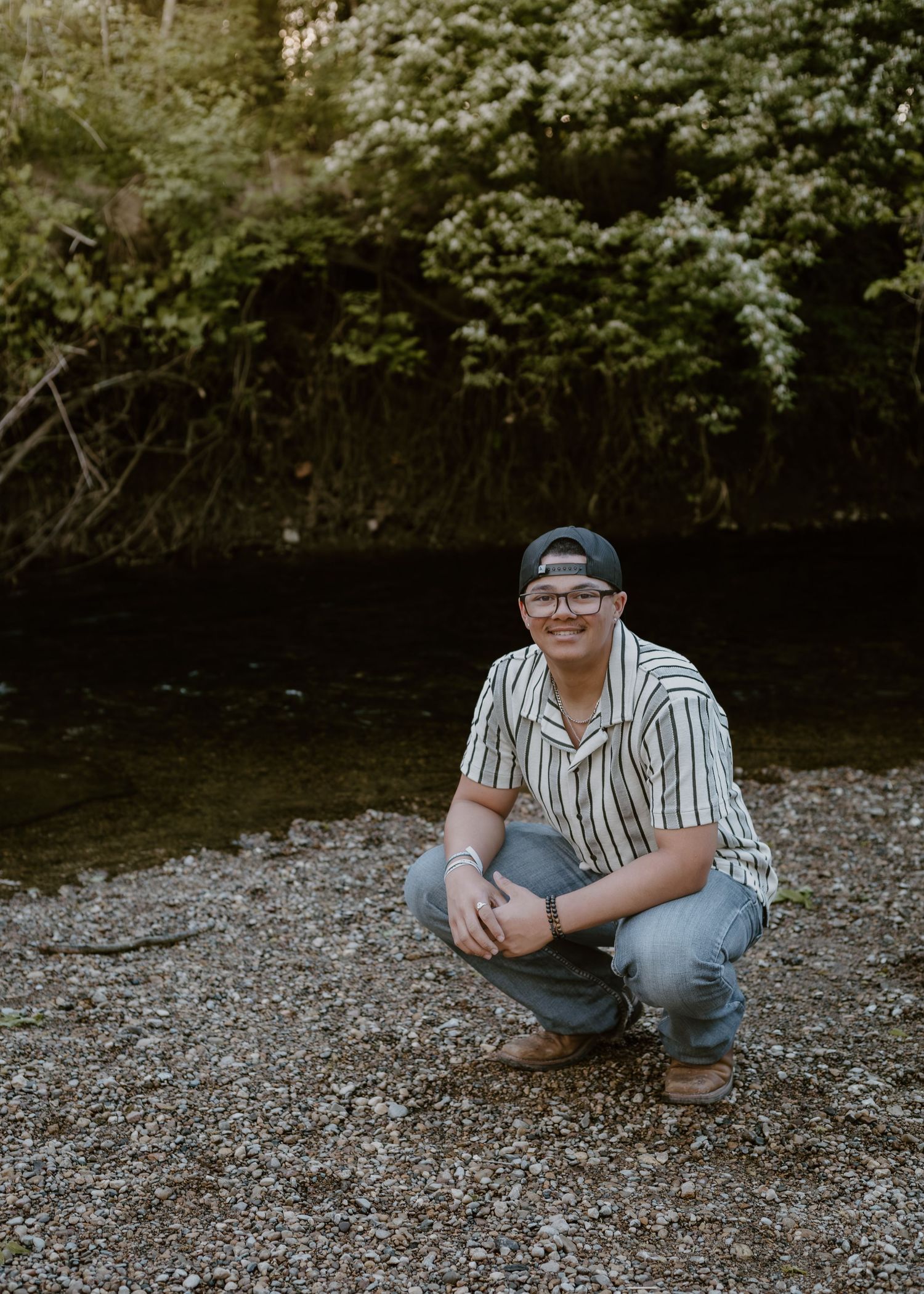 Smiling young man in striped shirt crouching by rocky creek shore with green trees behind him.