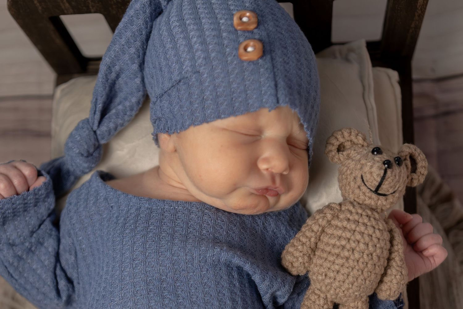 Sleeping newborn in blue knit outfit and hat cuddles a small crocheted teddy bear in a wooden chair.