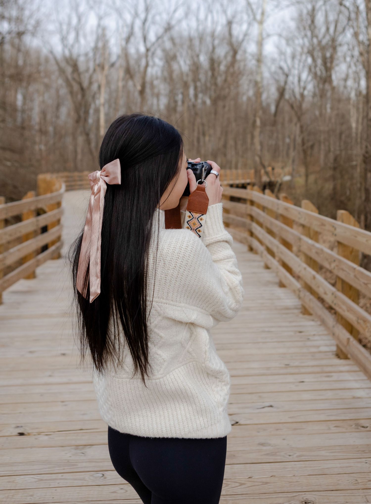 Young woman with black hair and pink bow photographs nature on a wooden bridge in winter woods.