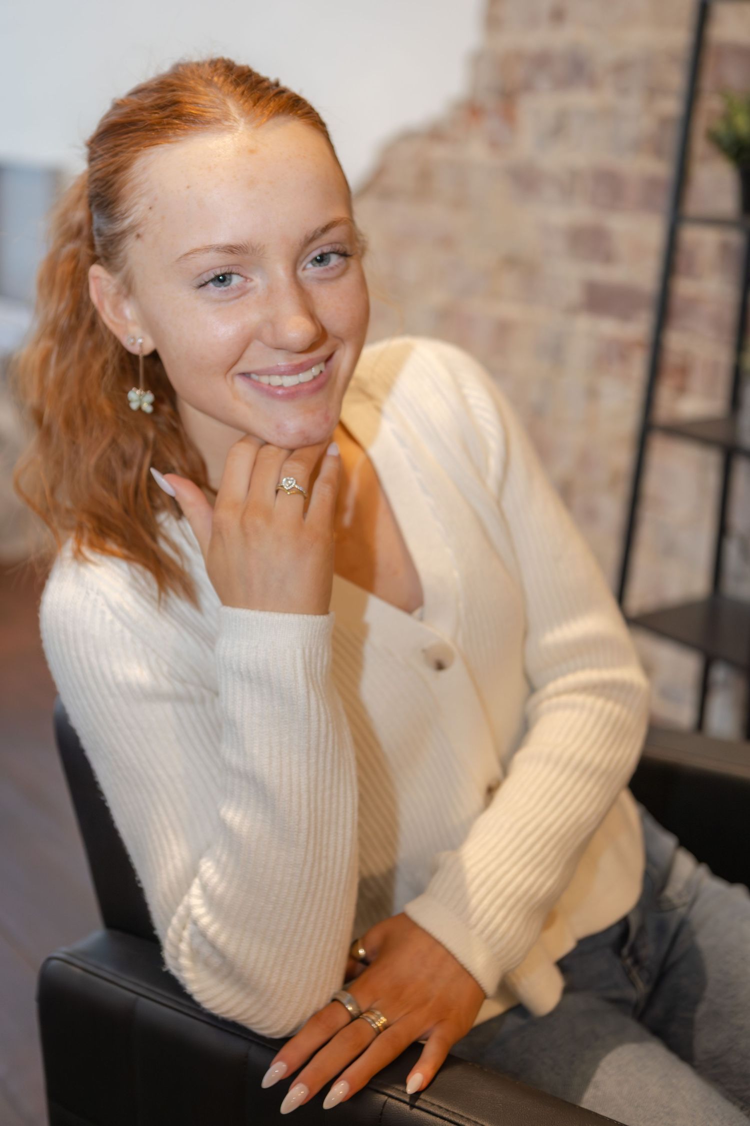 Smiling young redhead woman poses casually in a white cardigan in a modern salon interior.
