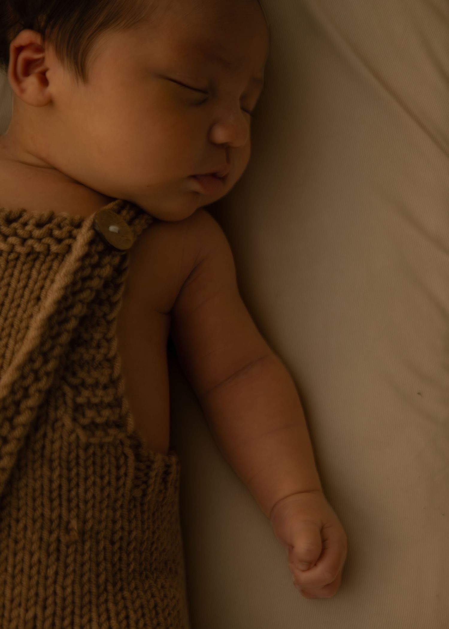 Sleeping newborn baby in a knit brown romper lying on a white surface.