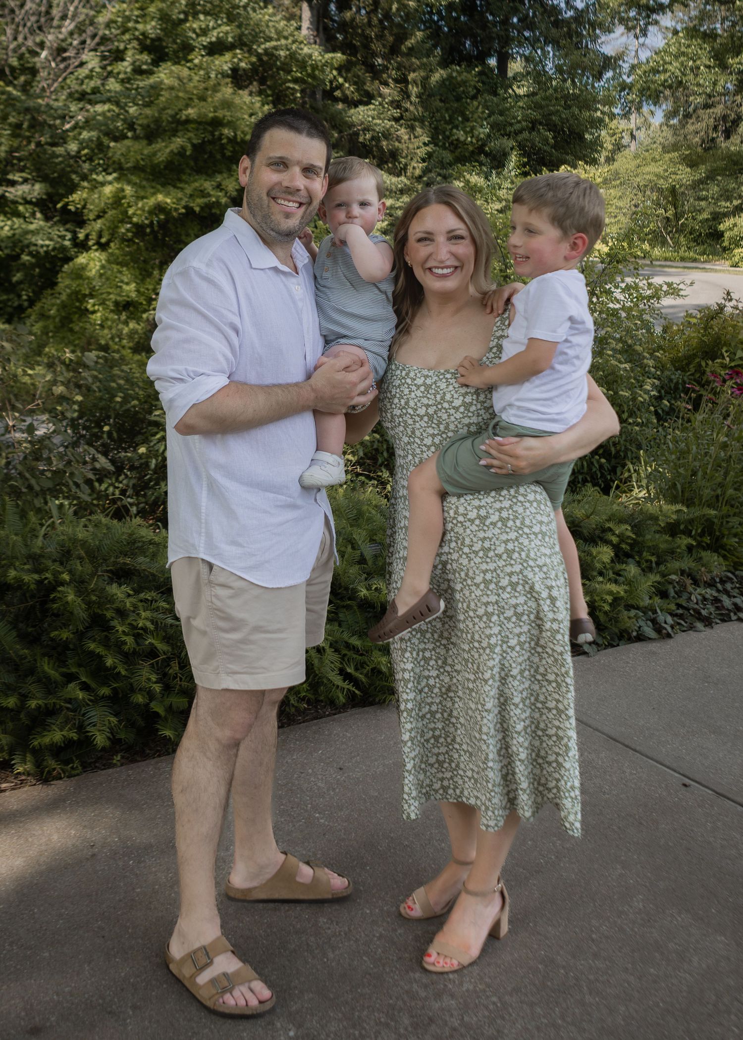 Happy family of four with two young children posing outdoors surrounded by lush green garden foliage.