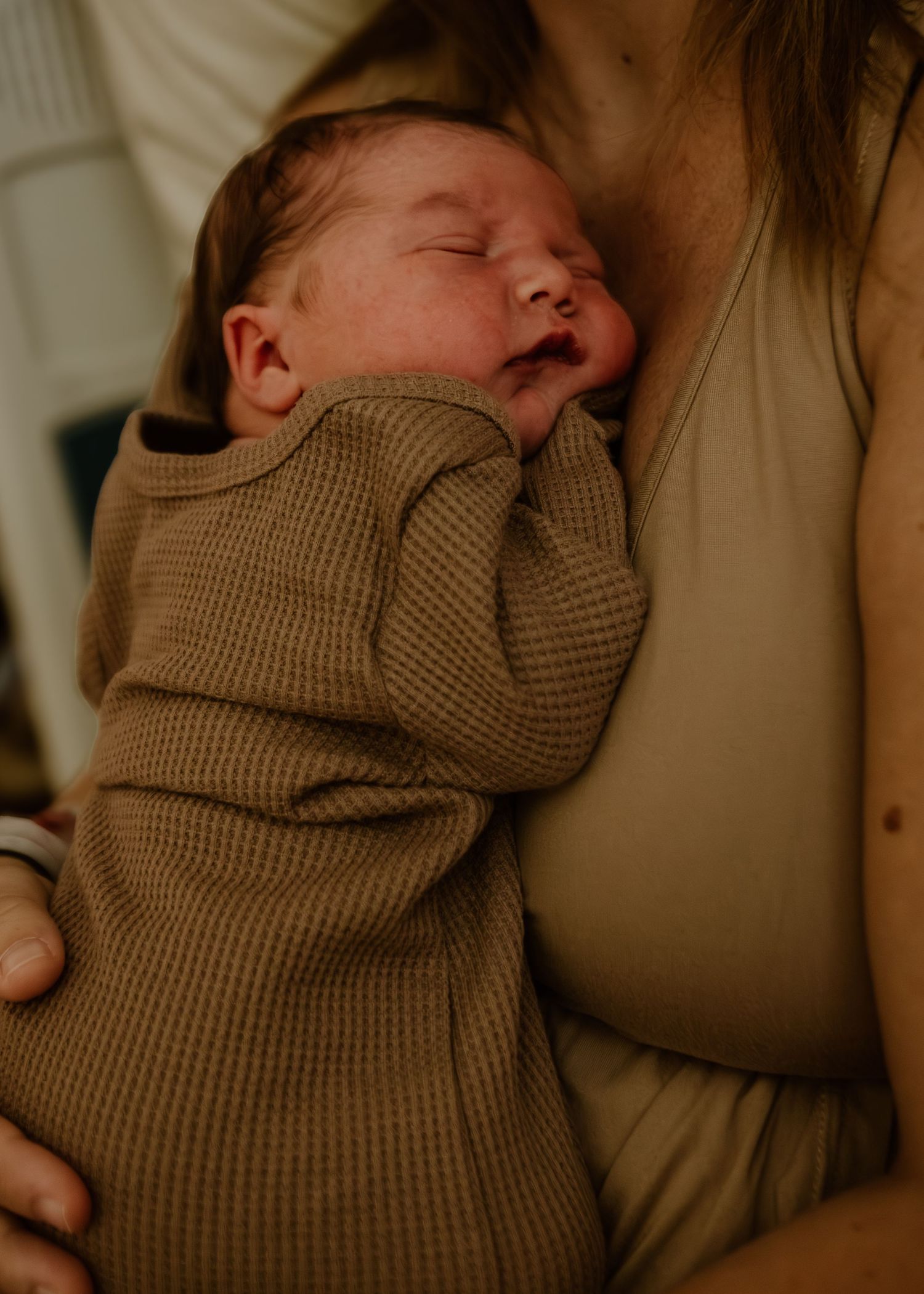Sleeping newborn baby in brown waffle knit outfit cradled in mothers arms.