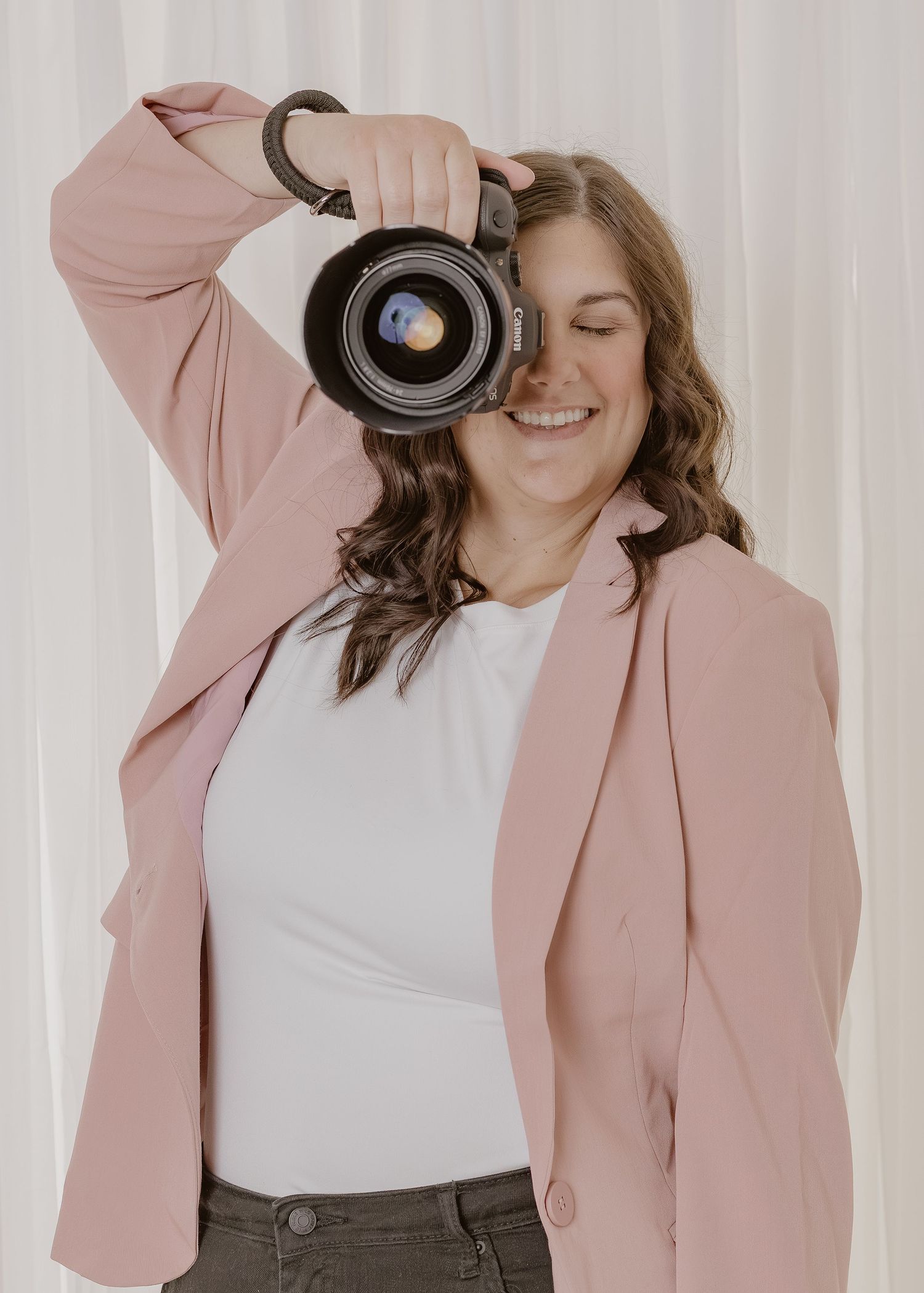 Smiling female photographer in pink blazer holds DSLR camera up to her face against white curtain backdrop.