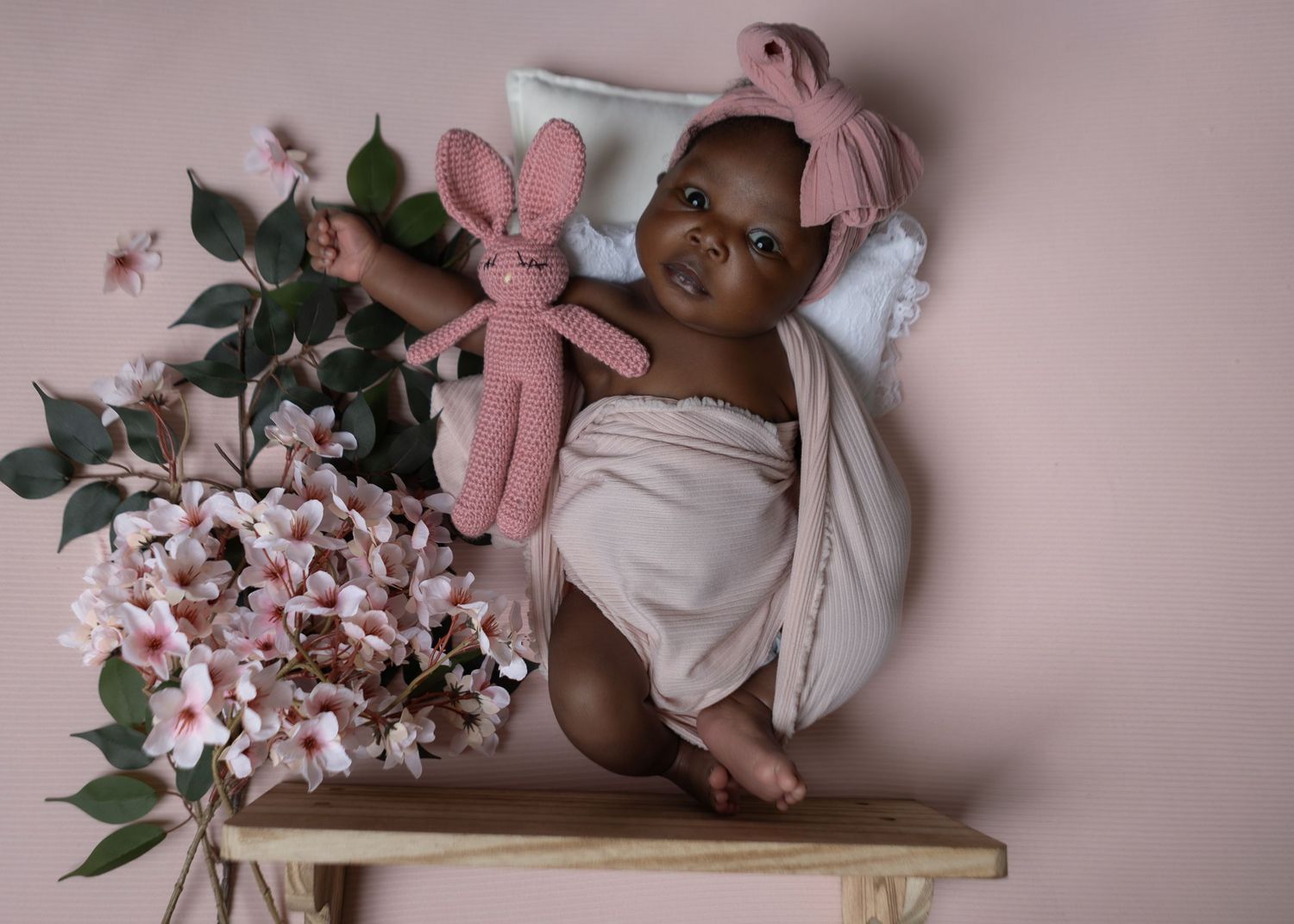 Adorable baby girl in a cream wrap sitting with pink flowers and a crochet bunny toy.