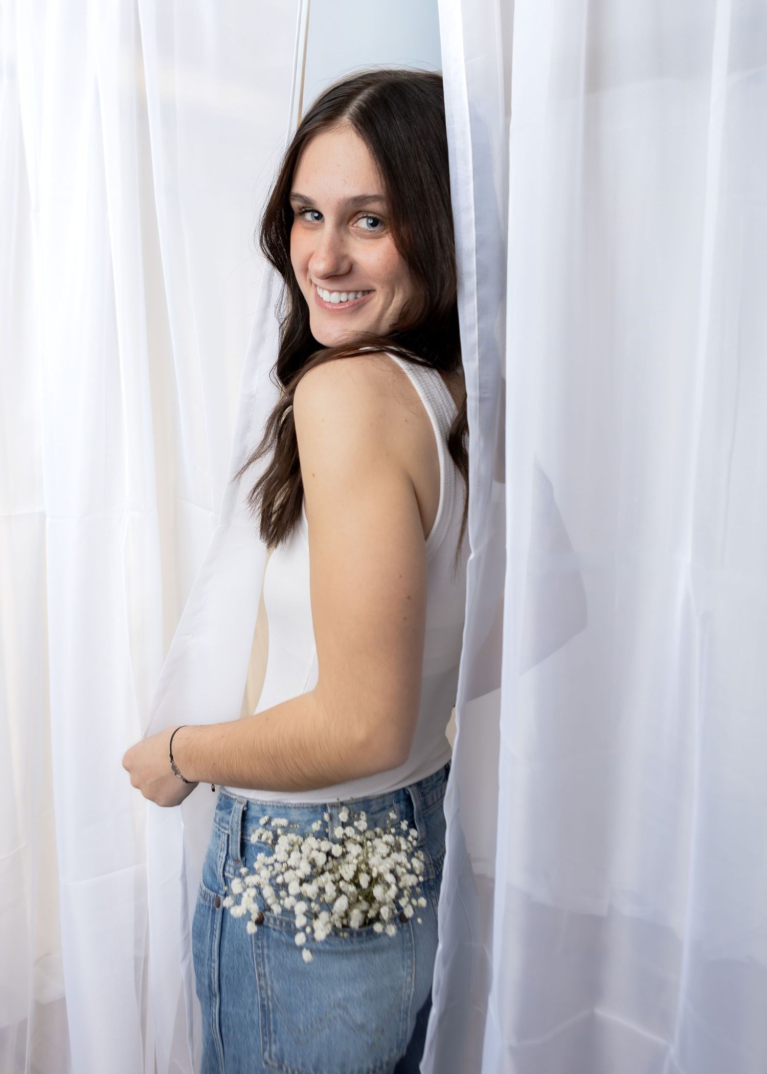 Smiling brunette teen girl posing between white sheer curtains with flowers tucked in her jeans.