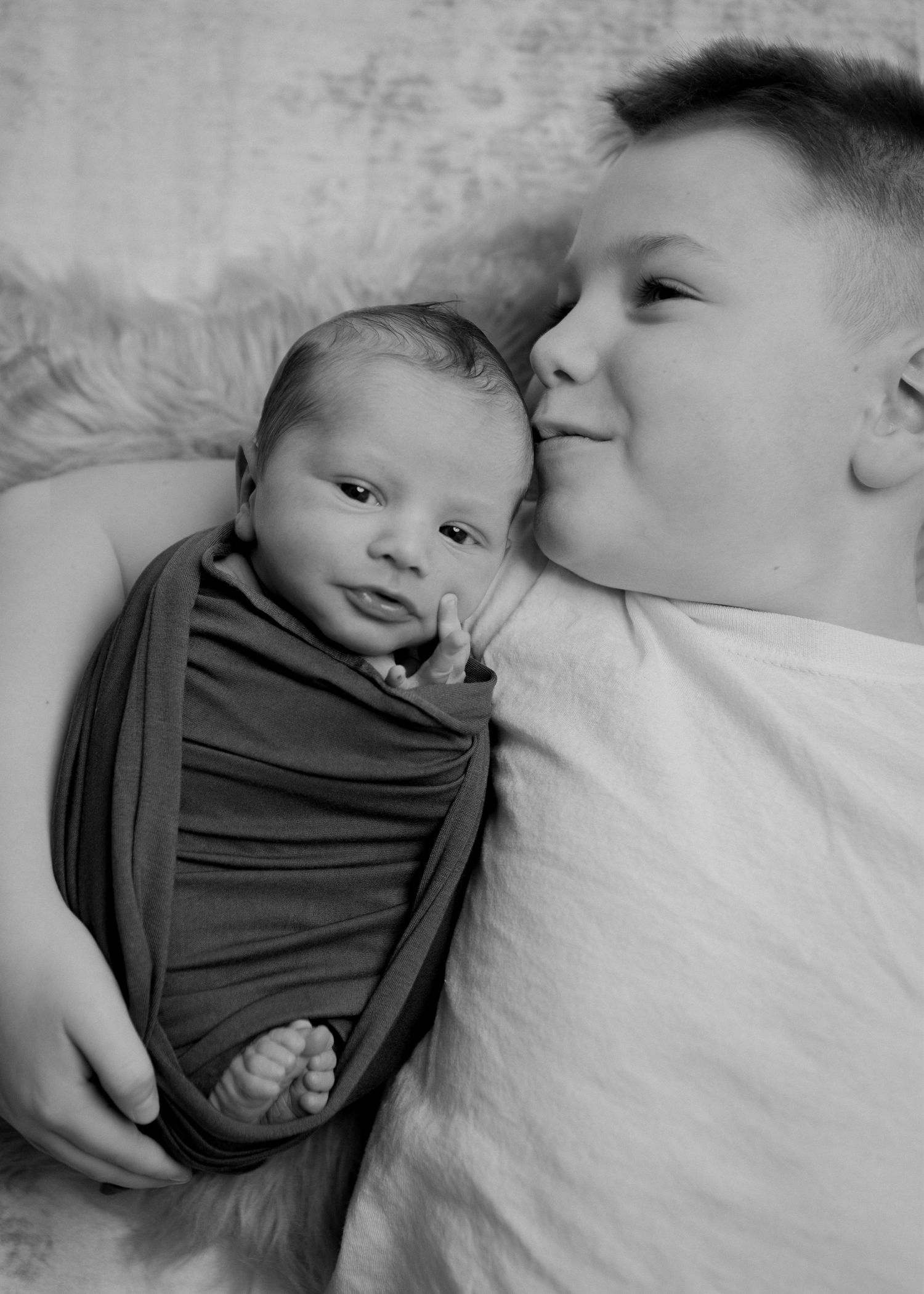 Black and white photo of older boy kissing newborn swaddled in wrap on fluffy backdrop.