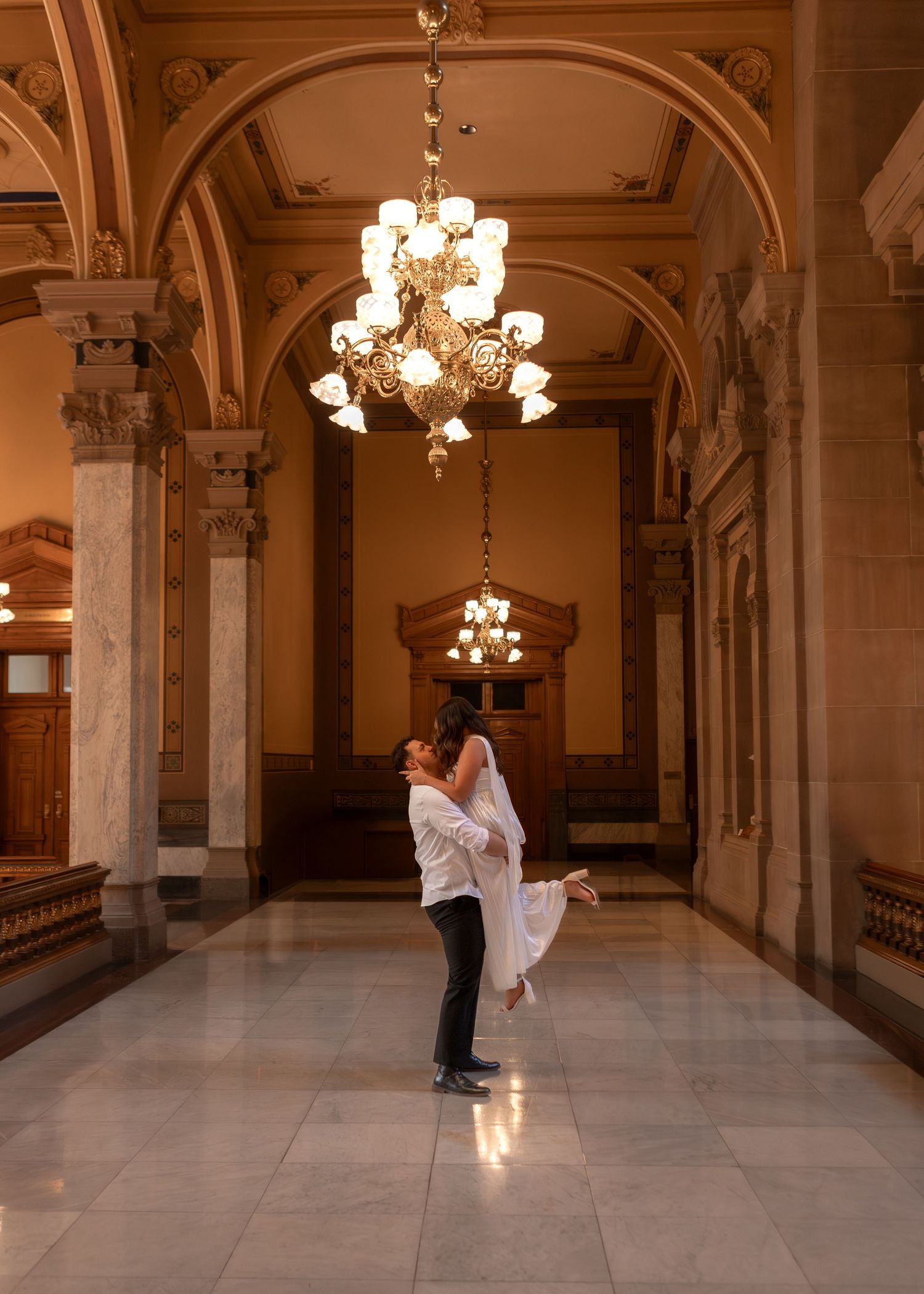 Groom lifting bride in elegant marble hallway with chandelier during wedding celebration.