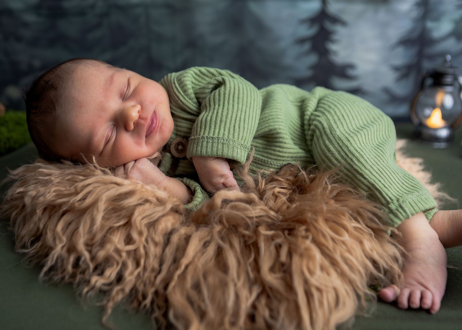 Sleeping newborn baby in a green knit outfit resting on a fluffy brown rug in a woodland setting.