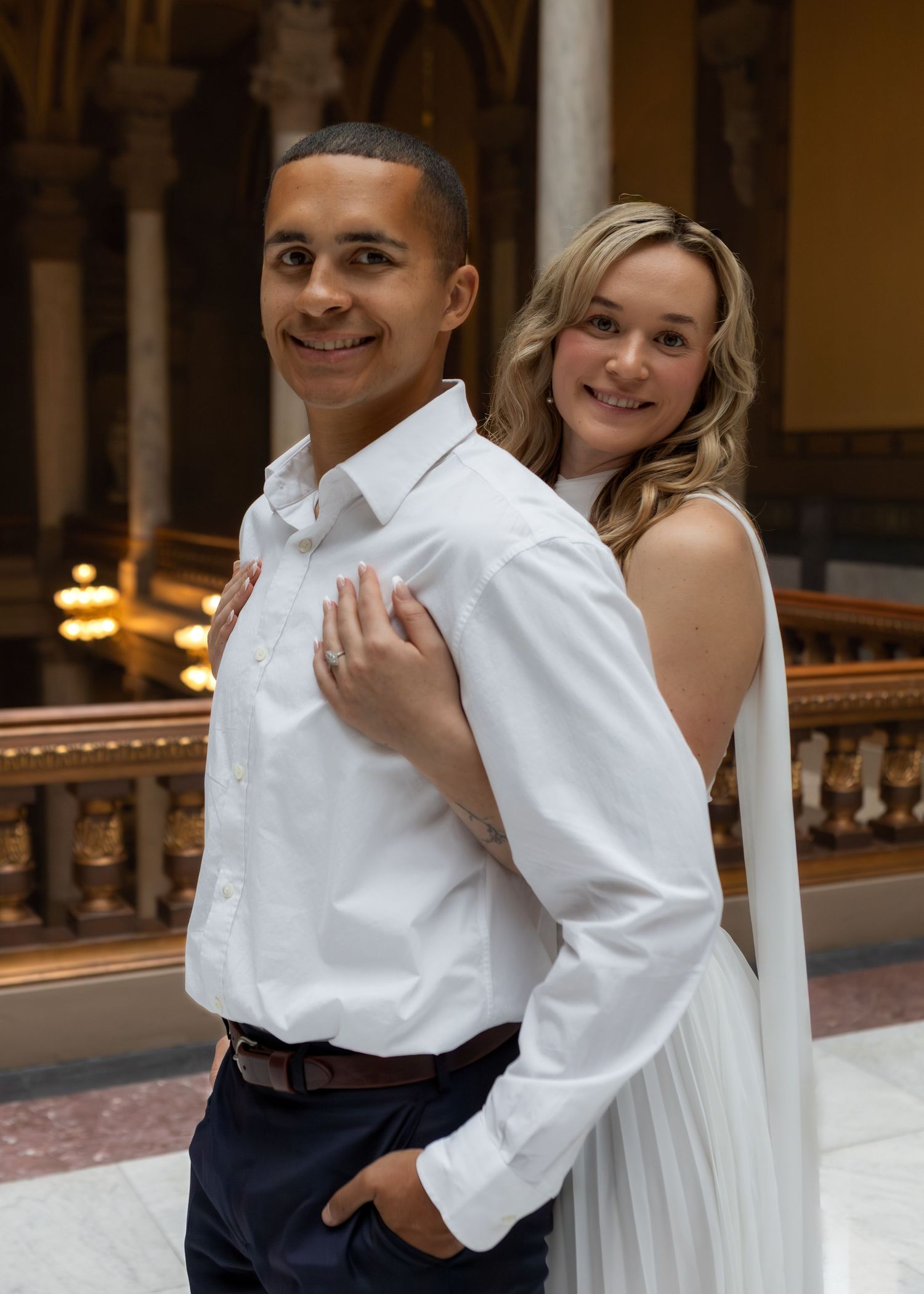 Couple in white wedding attire posing together in elegant indoor venue with ornate architecture.