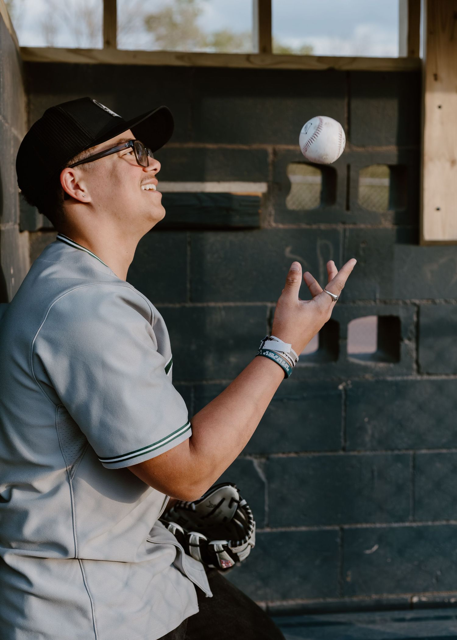 Young baseball player in gray jersey tossing a ball in the air while leaning against a brick wall outdoors.