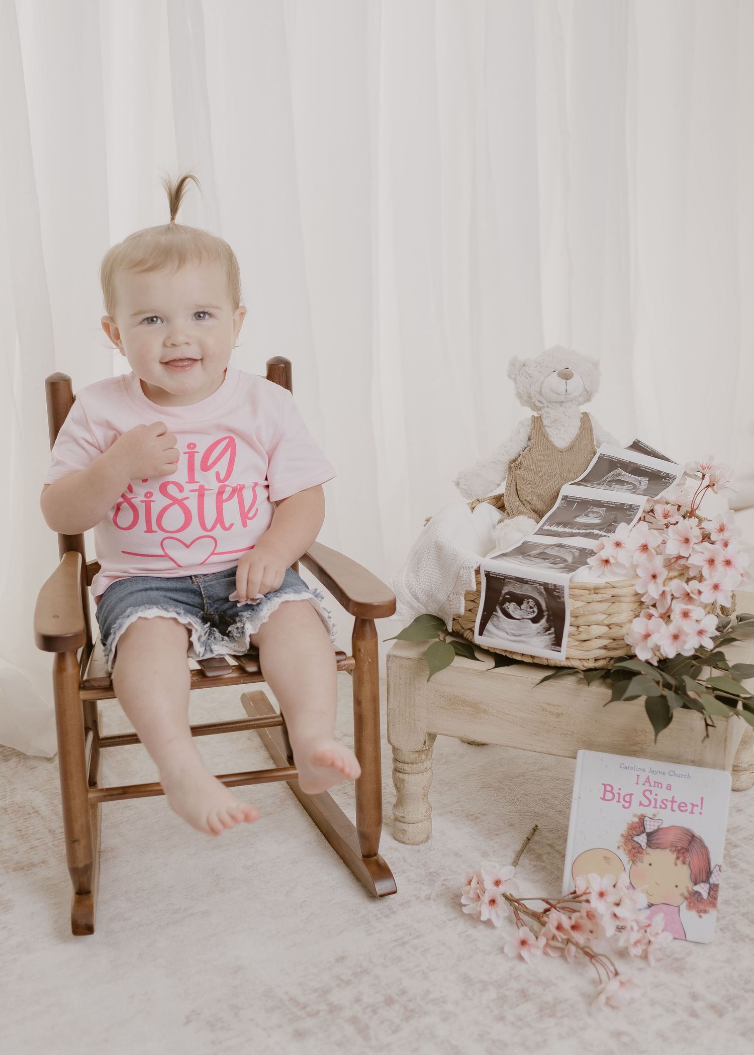 Smiling baby girl in Big Sister shirt sitting in wooden rocking chair beside ultrasound photos and flowers.