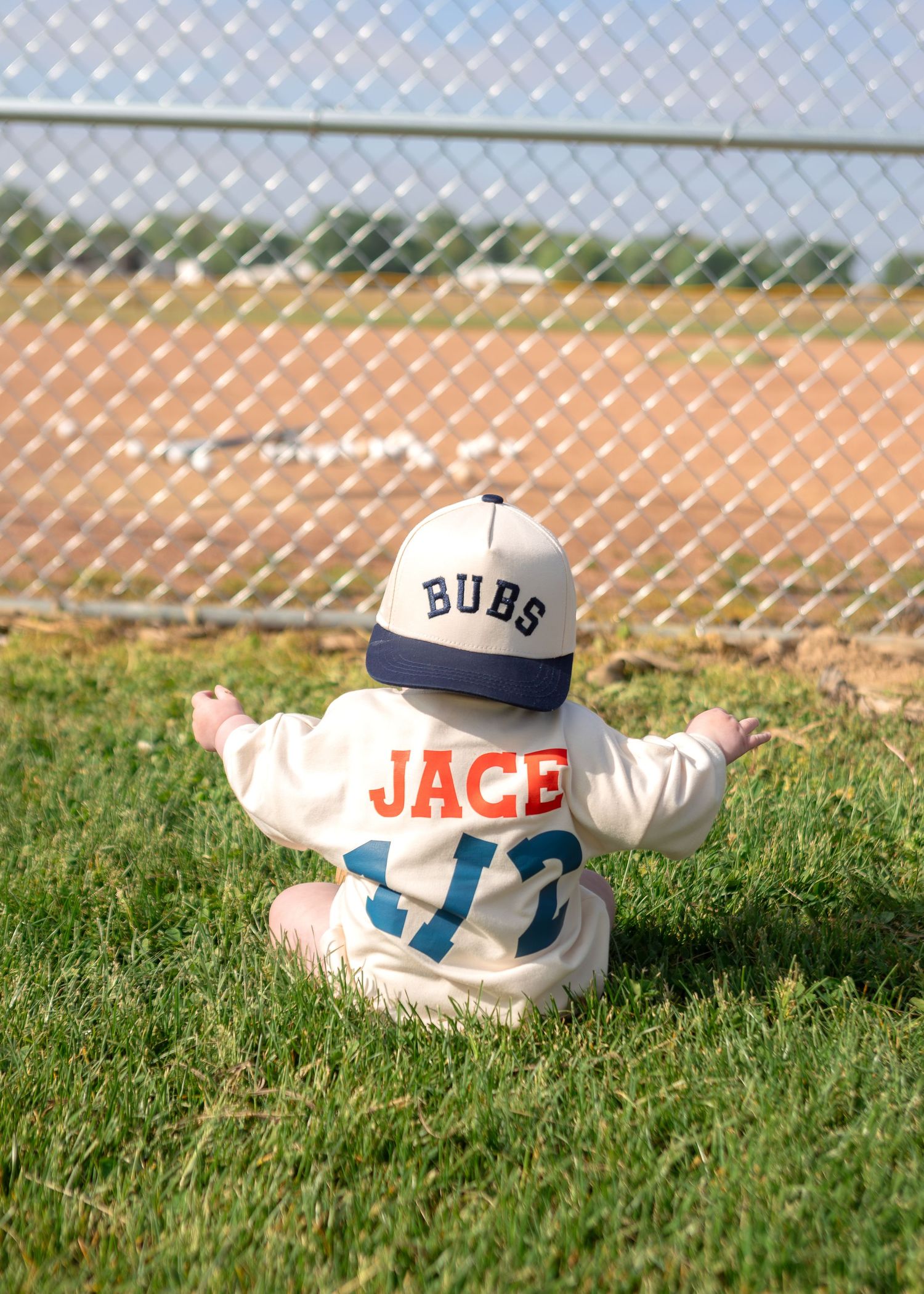 Baby named Jace in baseball outfit and Bubs cap sitting on grass by a ball field.