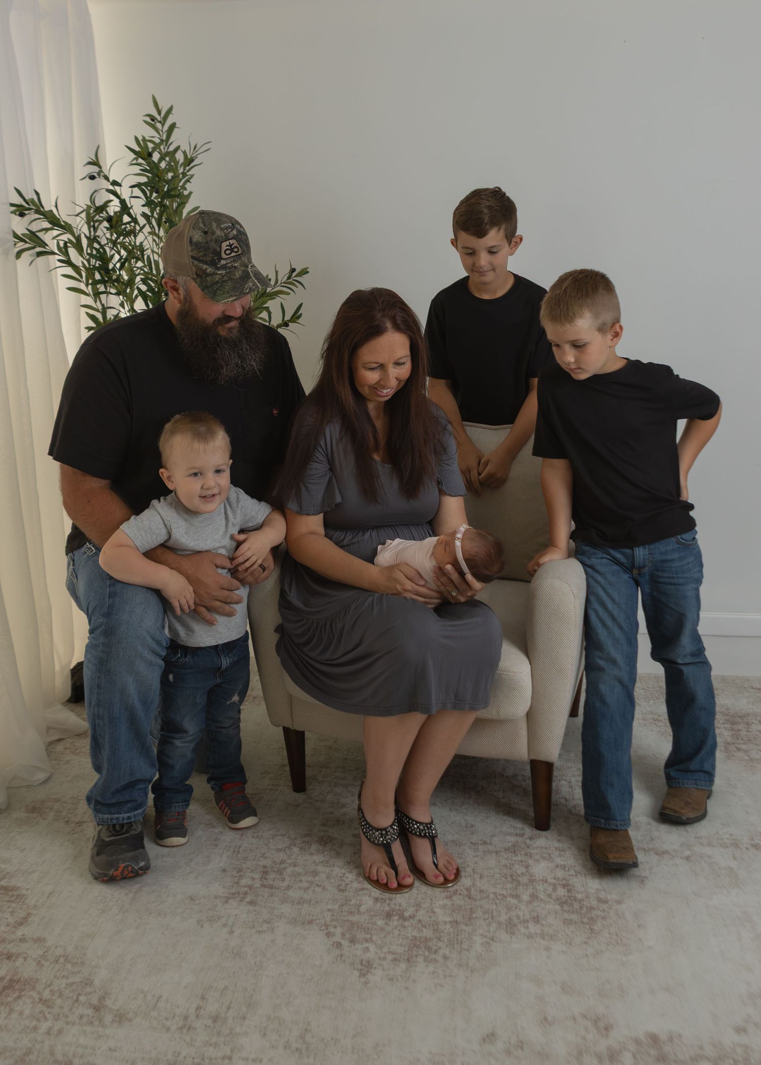 Family of six gathered around newborn baby in cozy white chair during indoor portrait session.