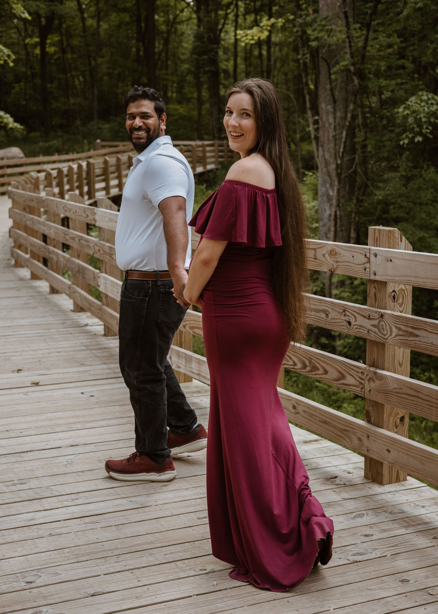 Couple holding hands on a wooden bridge, woman in elegant burgundy maternity gown.