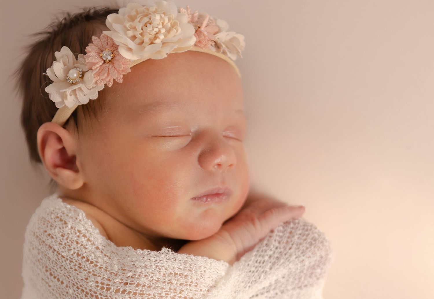 Sleeping newborn baby girl wearing a delicate floral headband, wrapped in a soft white knit blanket.