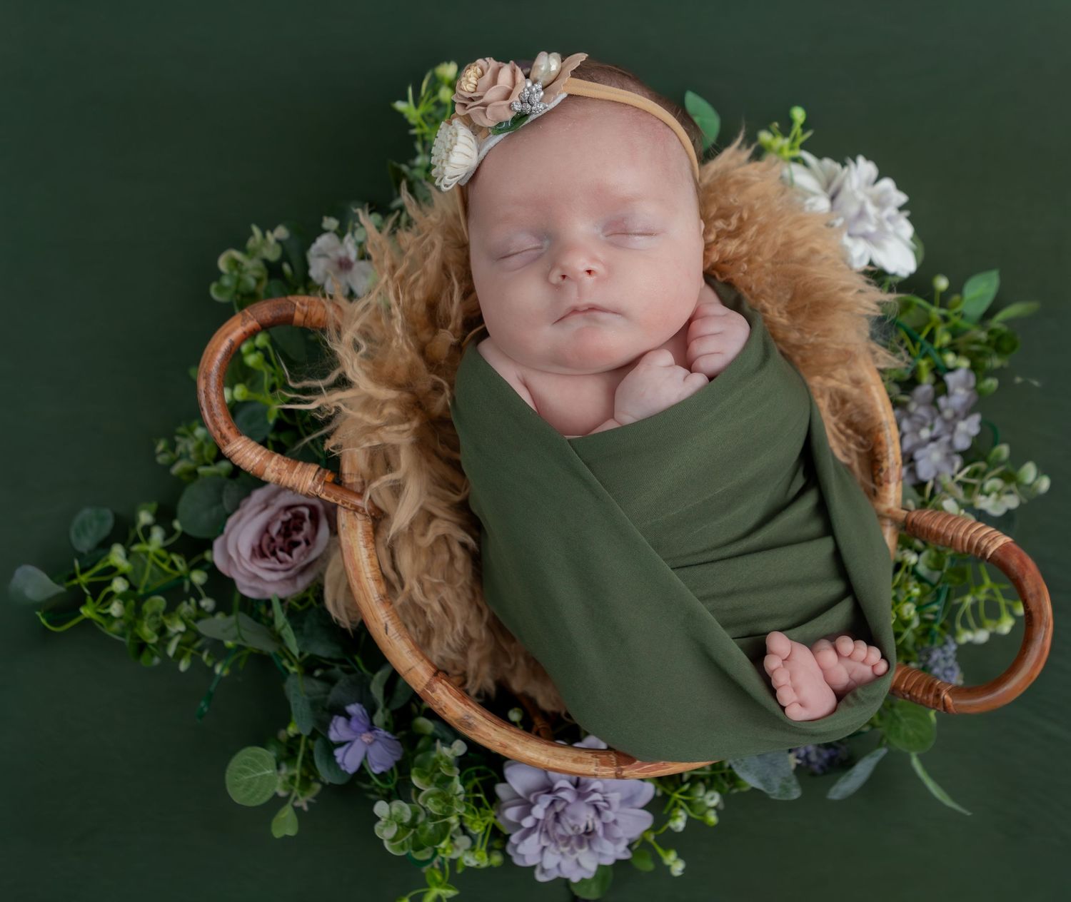 Sleeping newborn in green wrap with floral headband posed in basket with fresh flowers.
