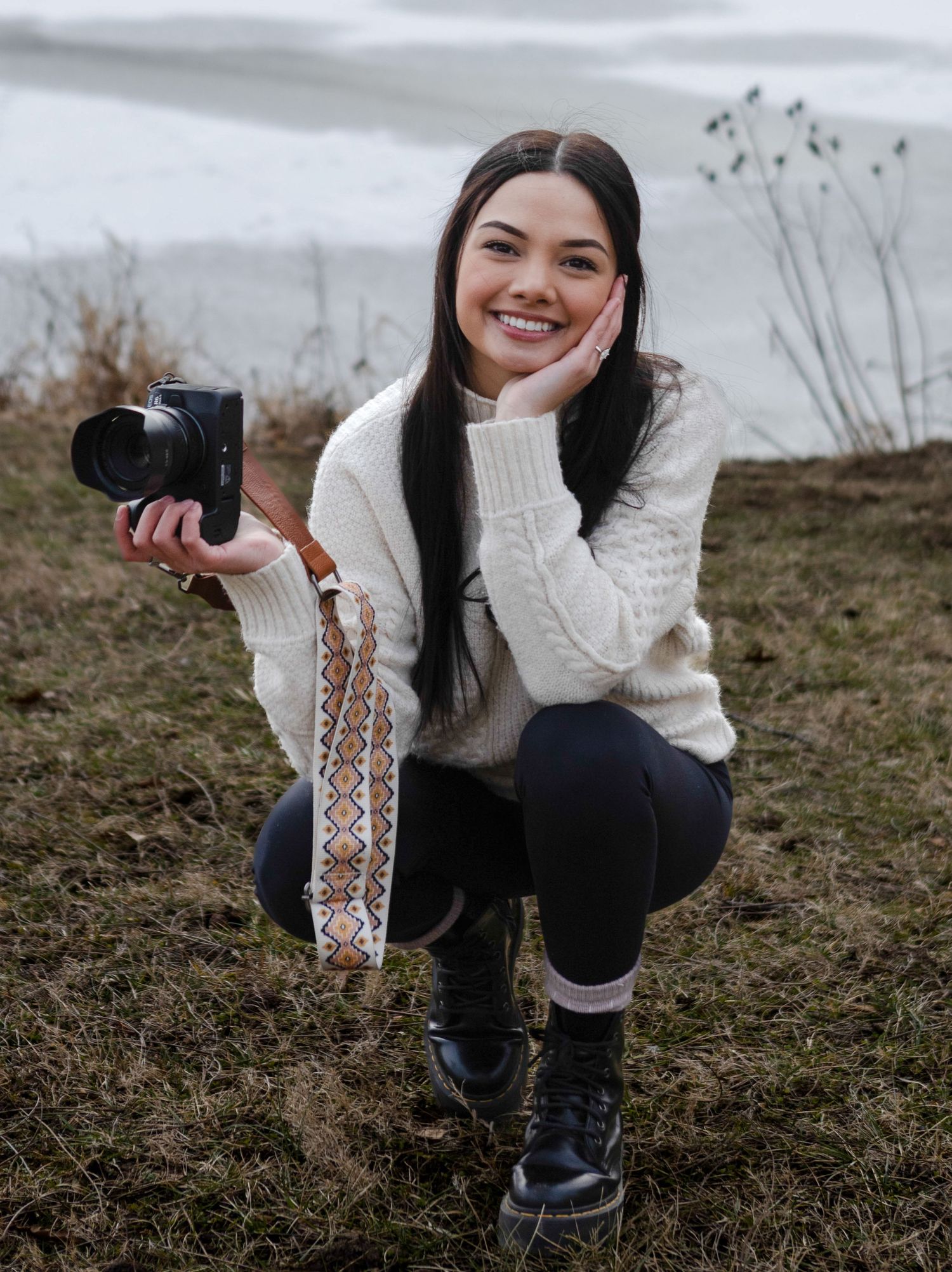 Smiling young woman crouching outdoors in winter, holding a camera and wearing a white knit sweater with black leggings.