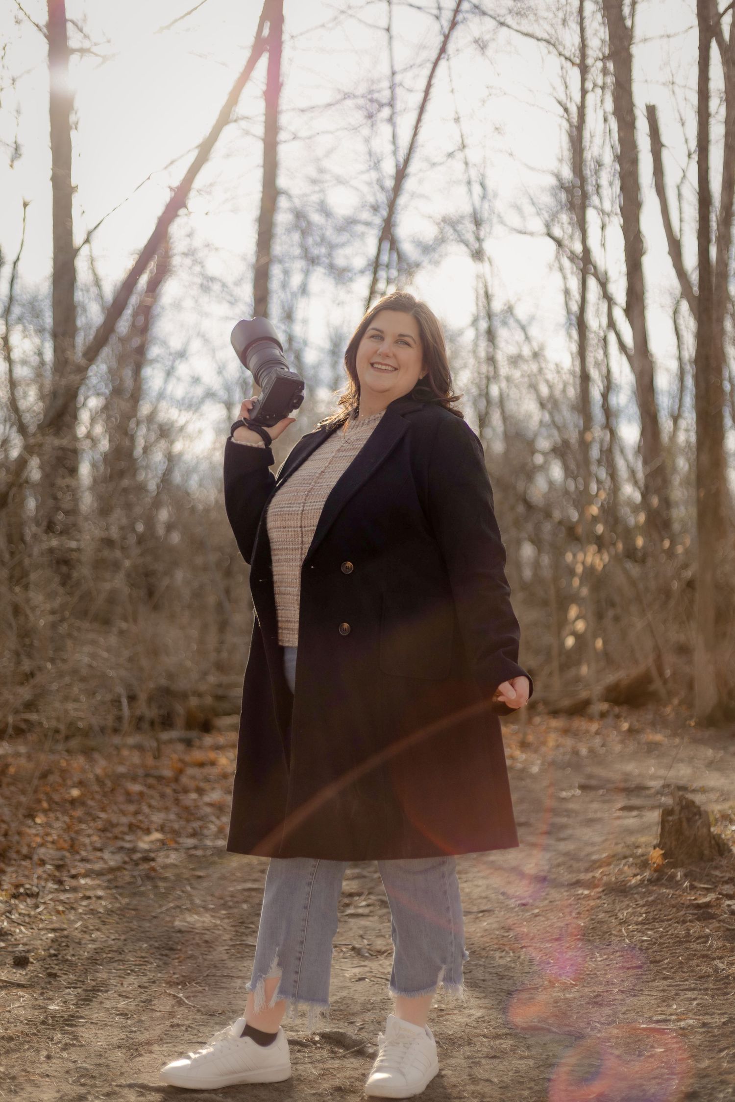 Smiling woman in black coat holding camera in sunlit bare winter forest with lens flare.
