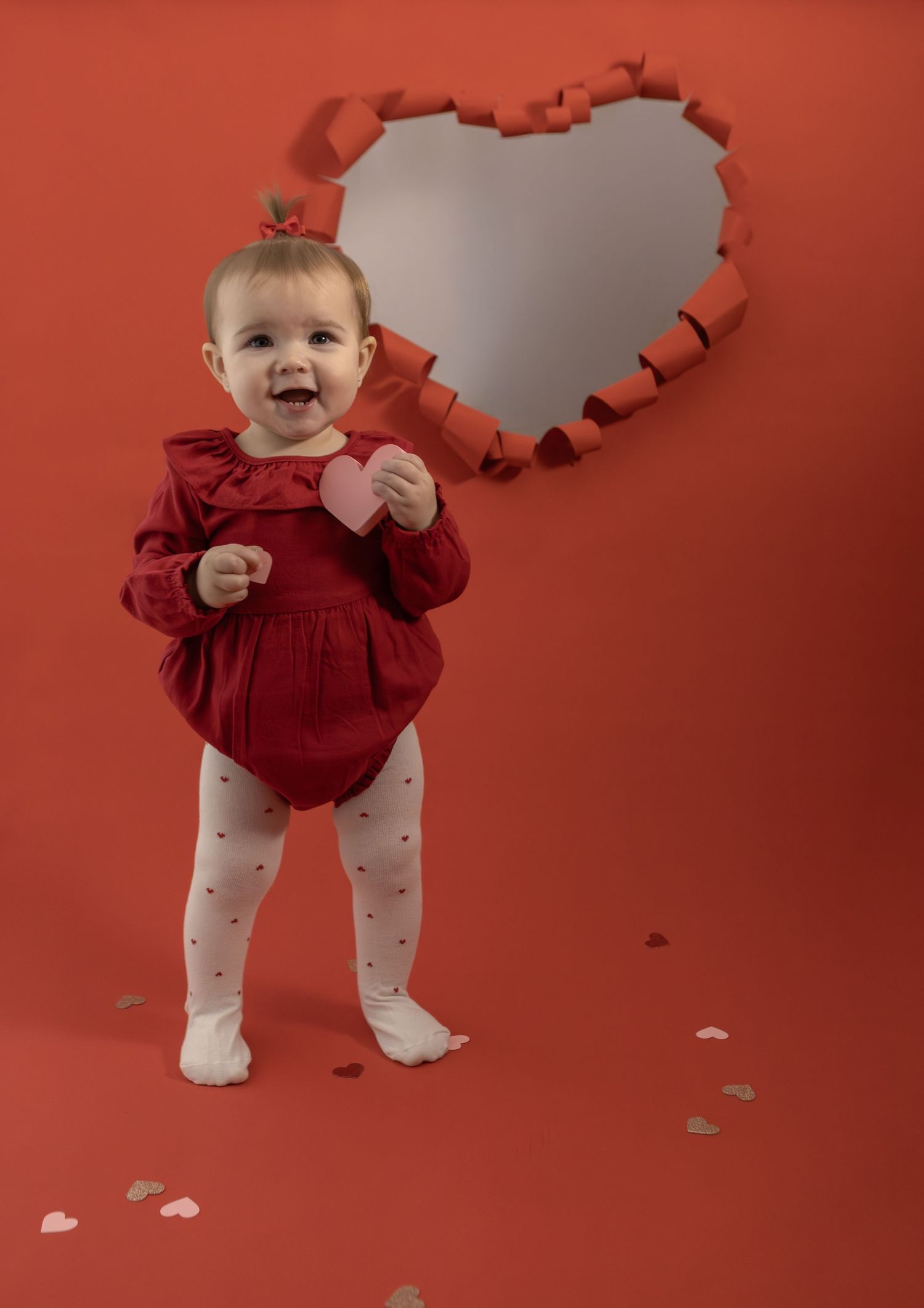 Smiling baby girl in red dress holds pink heart, standing before heart-shaped backdrop on Valentine's Day.