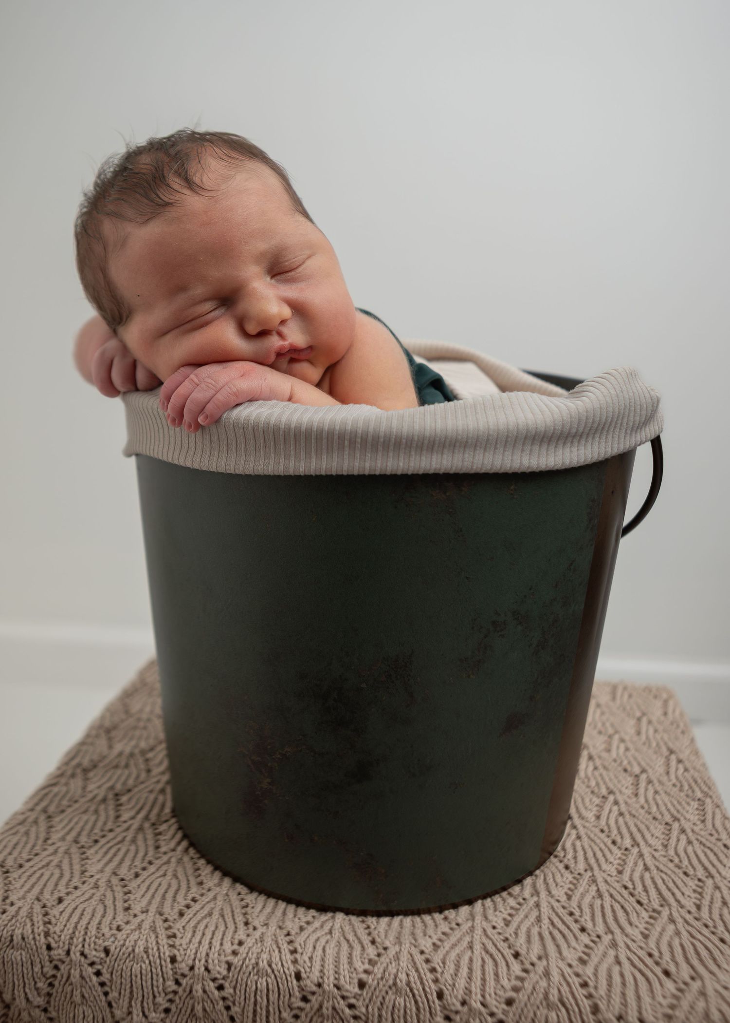 Sleeping newborn baby resting chin on hands inside a rustic green metal bucket prop.