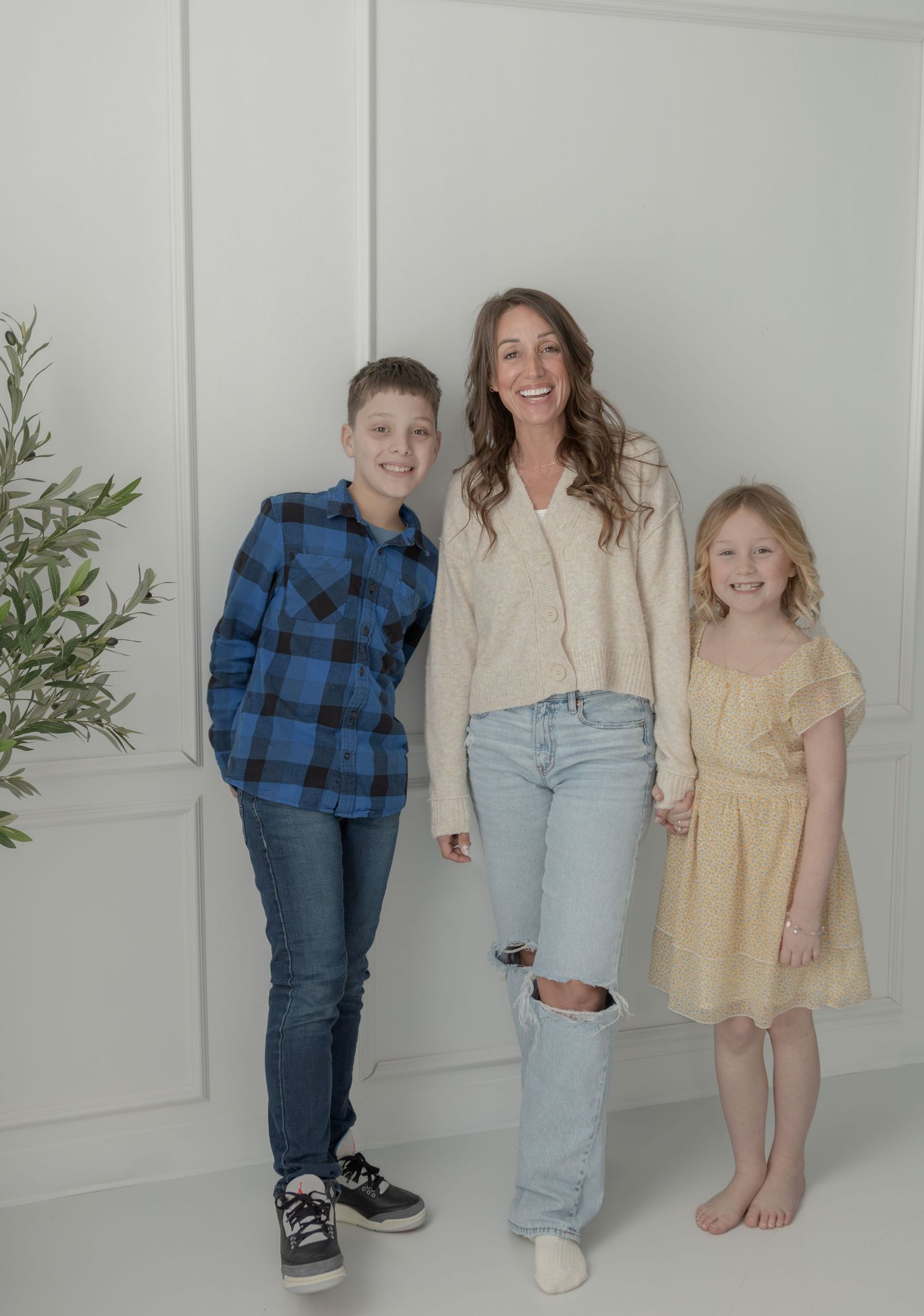 A smiling mom poses with her two children, a boy in a blue plaid shirt and a girl in a yellow dress, against a white wall.