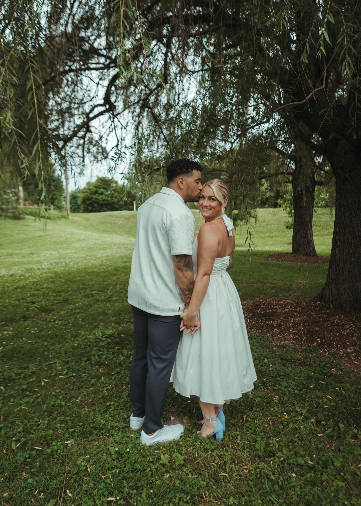 Couple embracing under a weeping willow tree outdoors, woman in white midi dress holding hands with tattooed man.