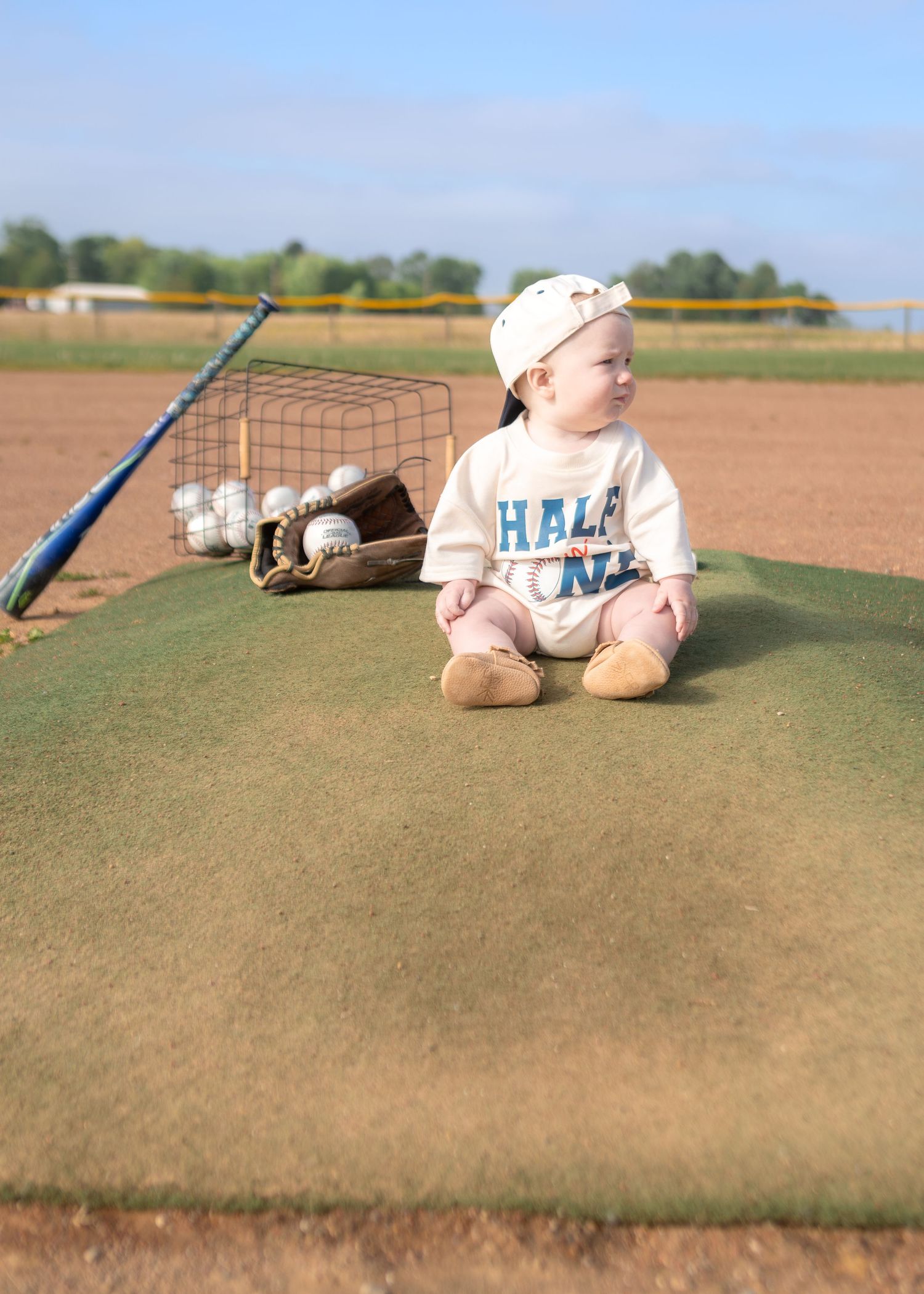 Baby in baseball cap and Half One onesie sits on field turf surrounded by bats, balls, and glove.