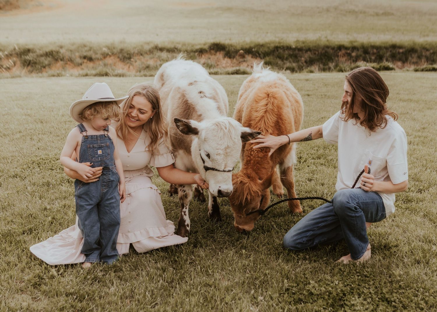 Two women and a child in overalls petting miniature cows on a sunny green farm.