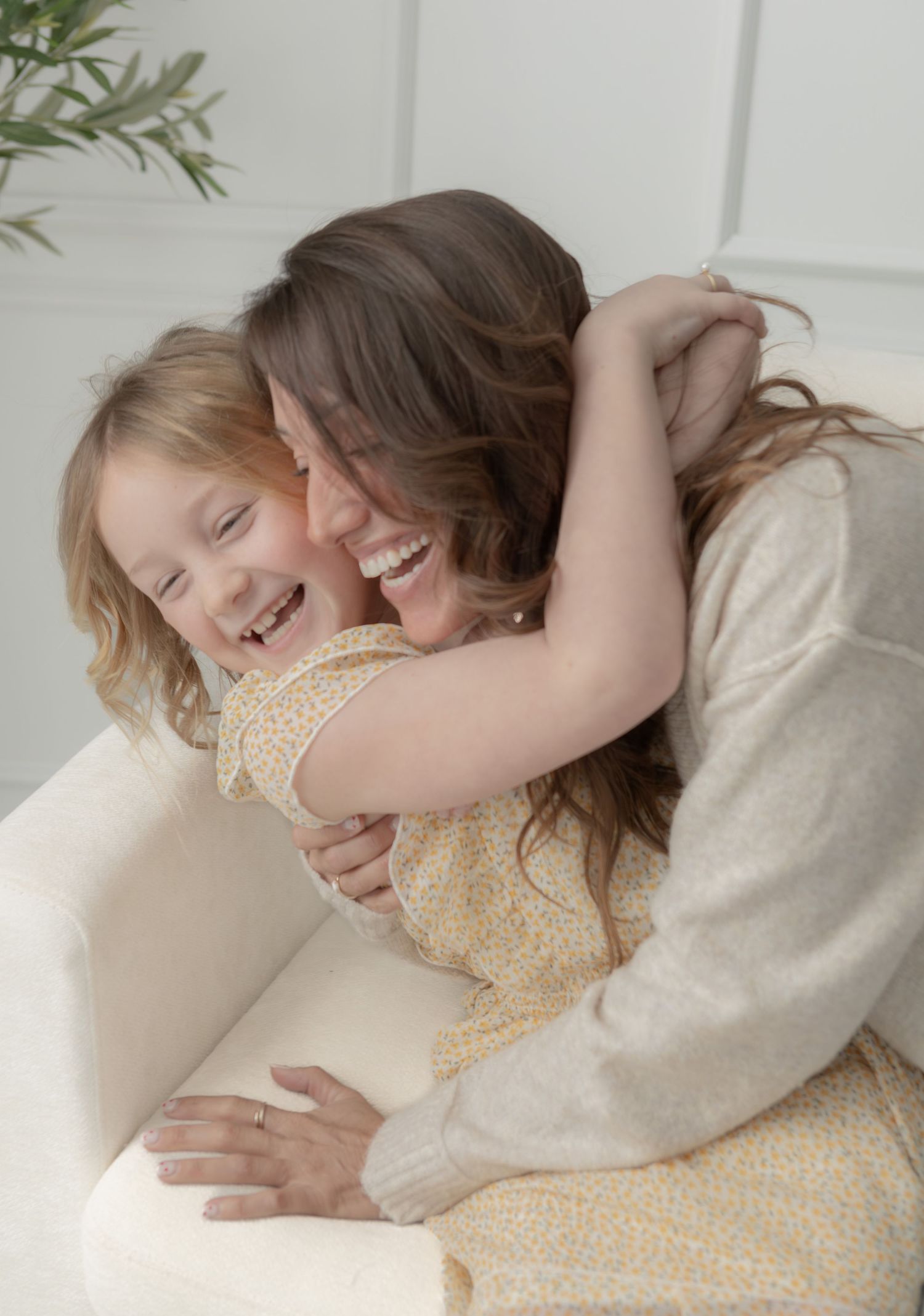 Laughing mother and blonde daughter share a joyful hug on a white couch in a bright room.