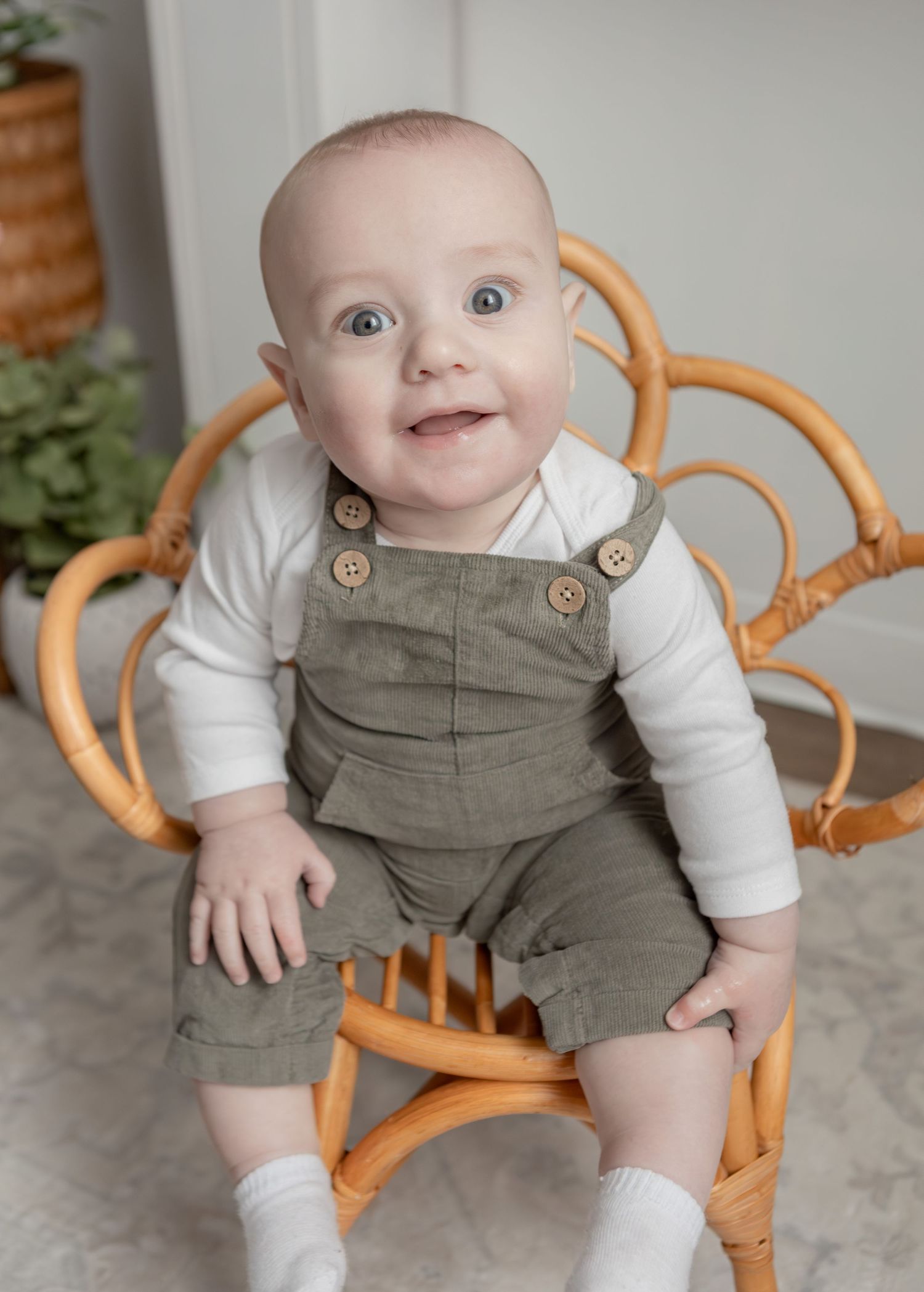 Adorable blue-eyed baby boy sitting in rattan chair wearing olive green overalls and white long sleeve shirt.