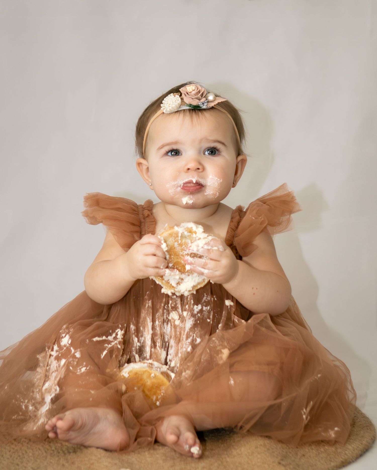 Baby girl in brown tulle dress with floral headband enjoying her first birthday cake smash.