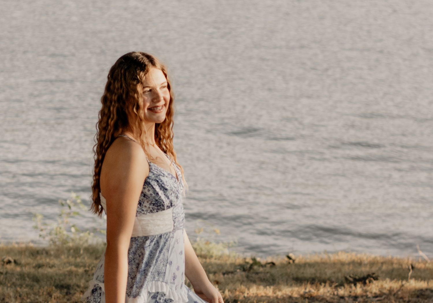 Young woman with curly red hair smiling in a floral sundress near a lake on a sunny day.