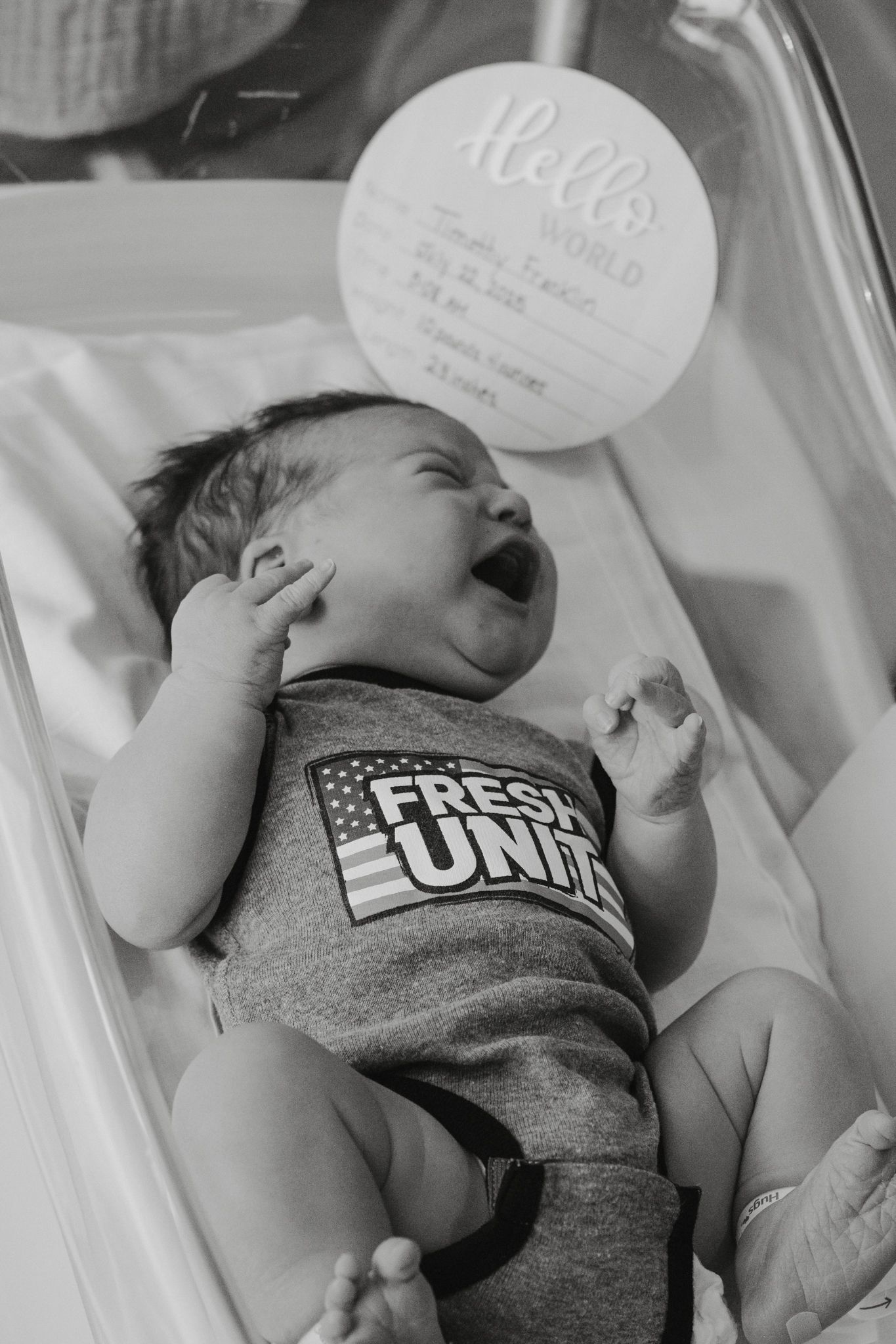 Black and white photo of a yawning newborn baby in a Fresh United onesie lying in a hospital bassinet.