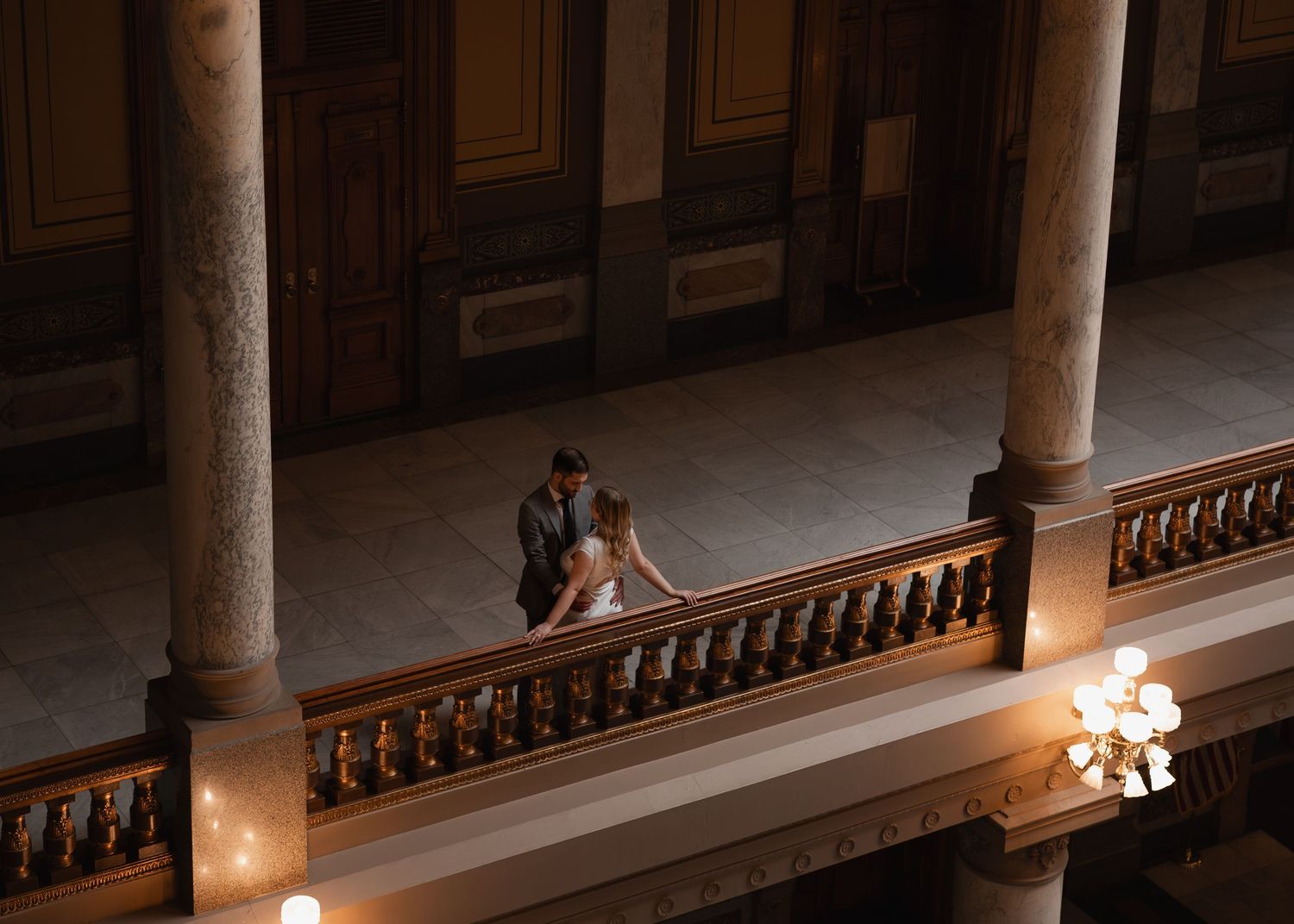 A couple shares a romantic moment on a grand balcony inside a stunning classical architecture building.