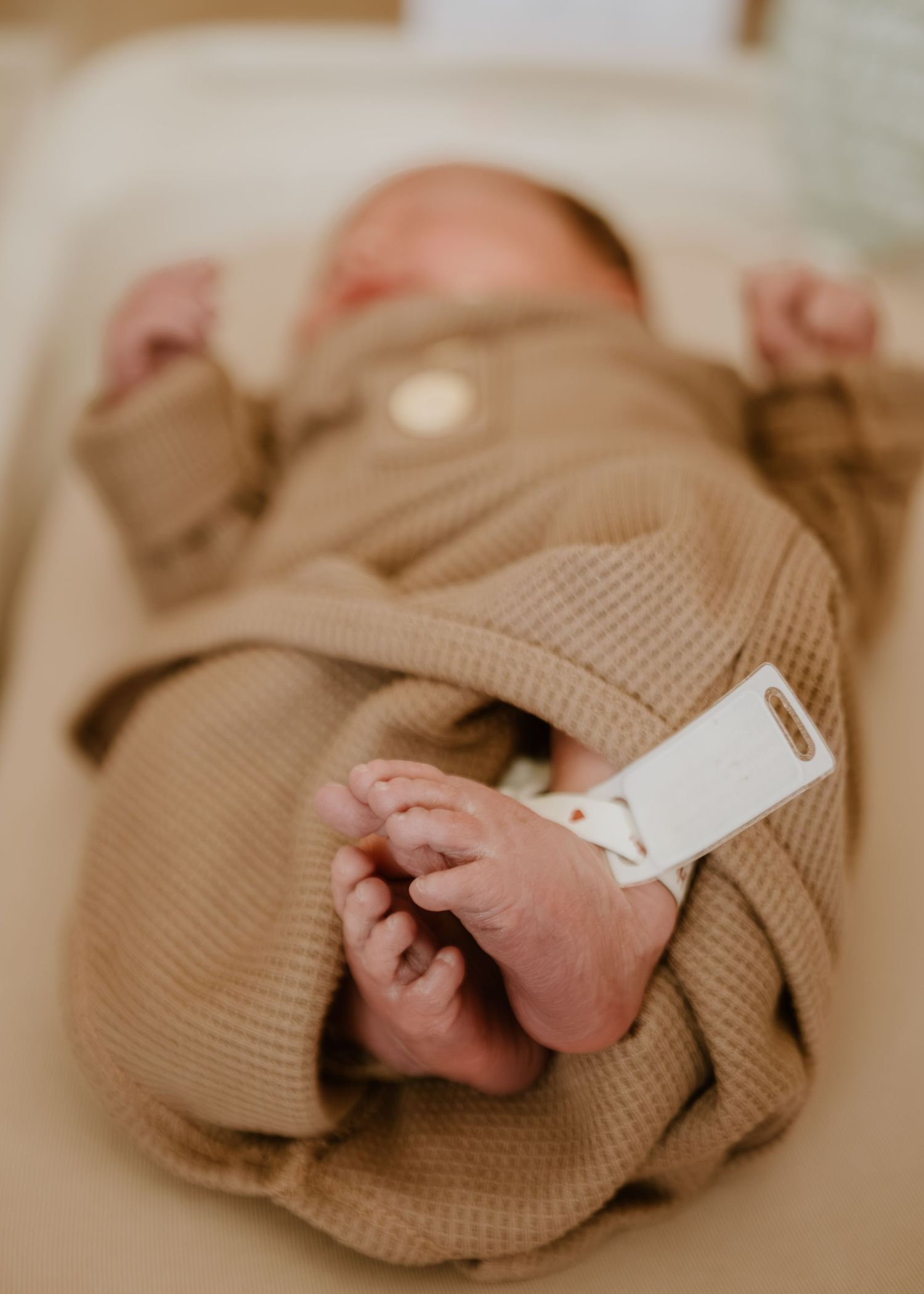 Close-up of newborn baby feet in a tan knit swaddle with hospital ID tag in a bassinet.