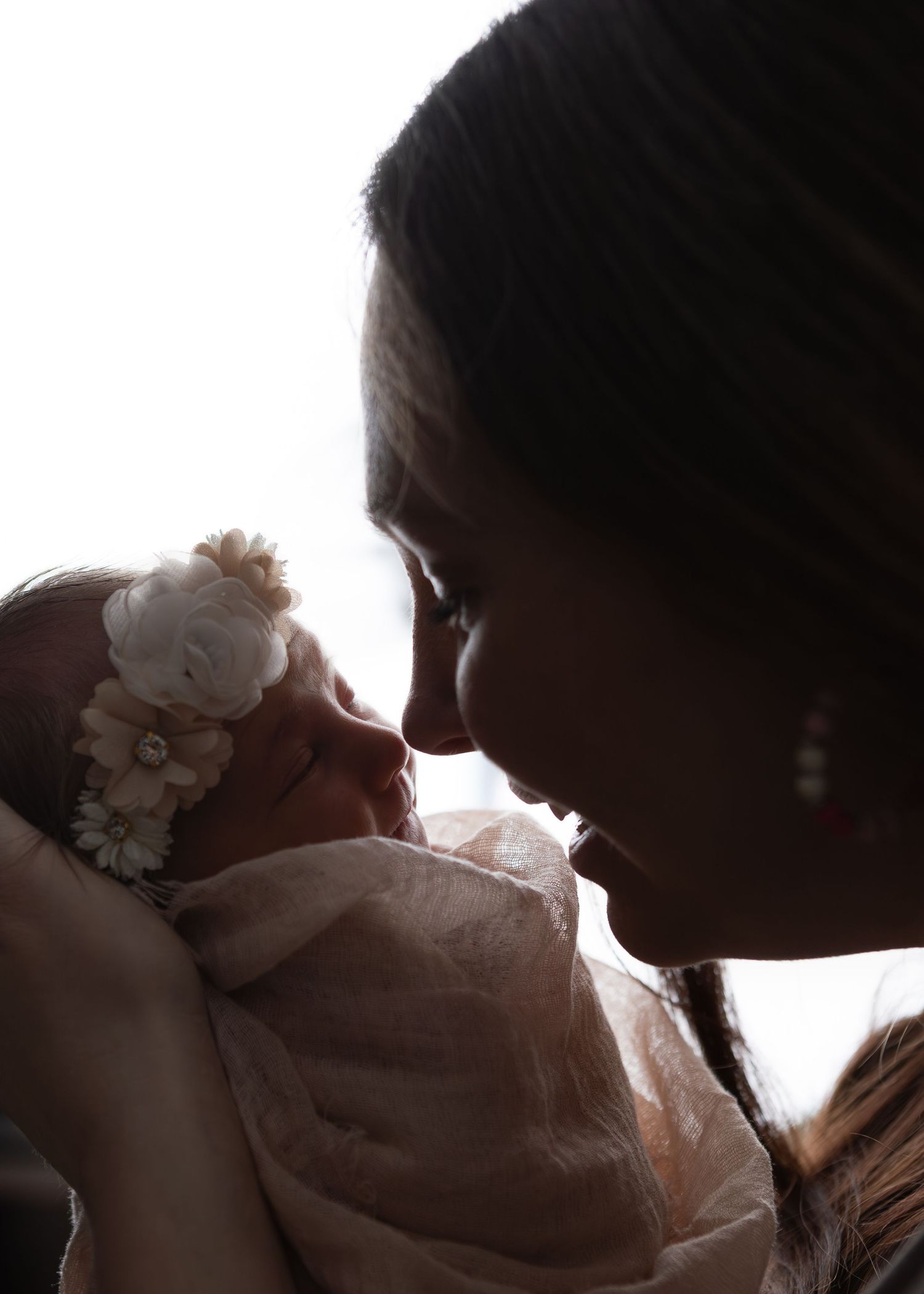 Tender silhouette of a mother gazing at her flower-crowned newborn baby in soft backlight.
