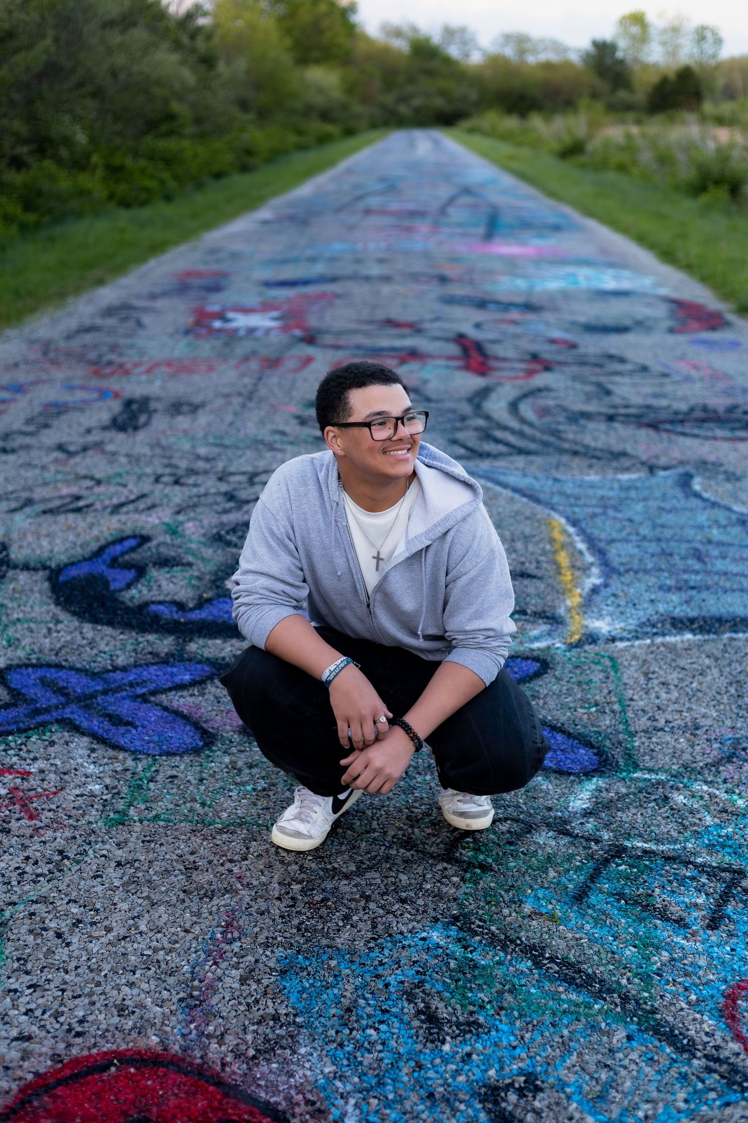 Smiling teen in gray hoodie crouching on colorful graffiti-covered pavement path surrounded by green trees.