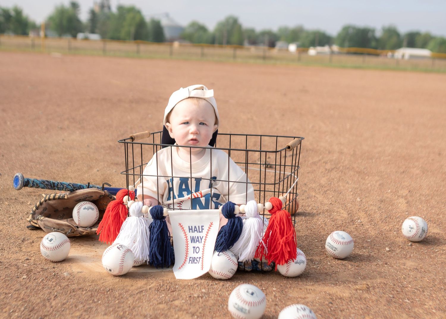 Baby named Jace sitting in a wire basket surrounded by baseballs on a dirt field.