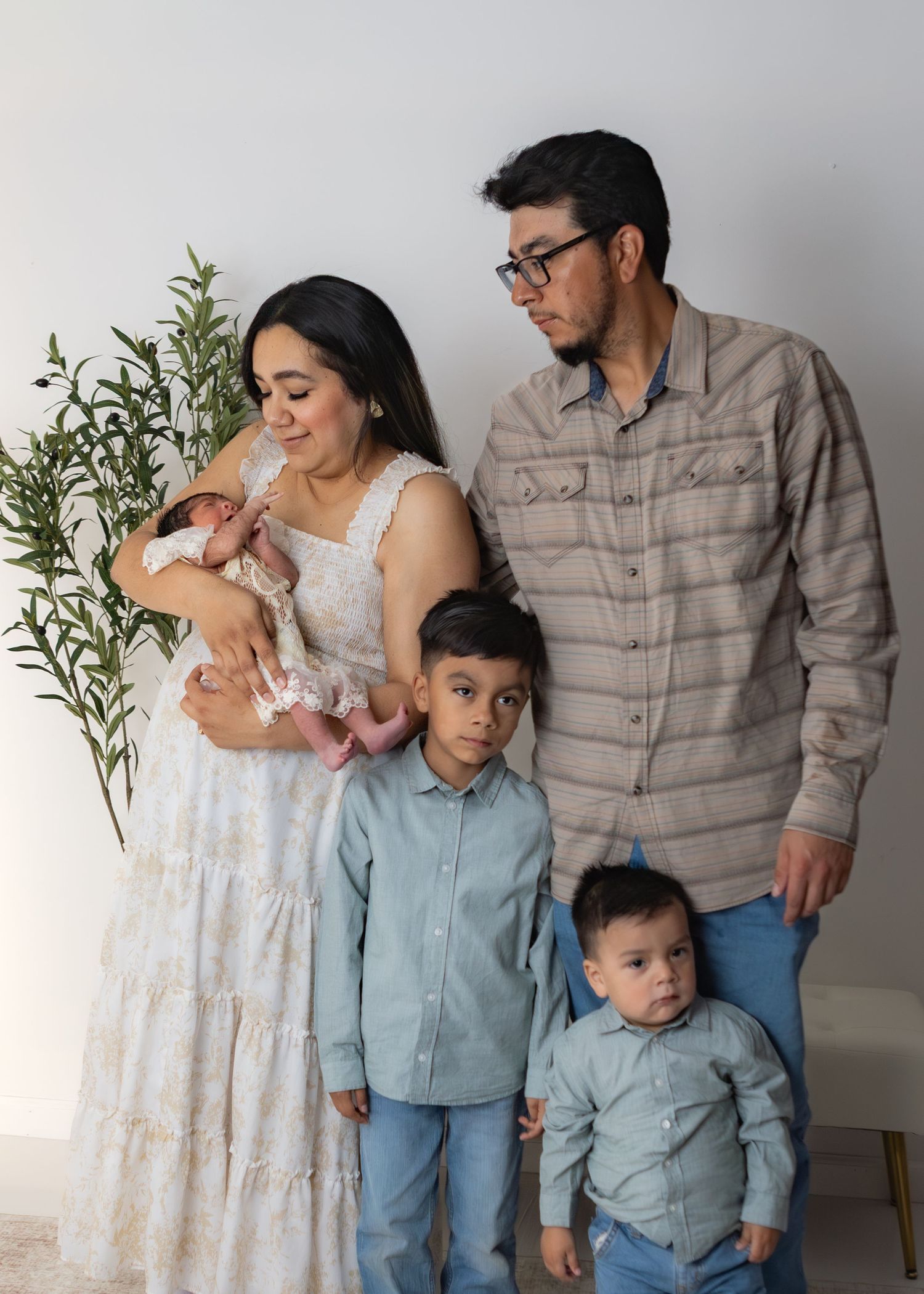 Family of five with newborn baby girl poses beside greenery in a bright white studio setting.
