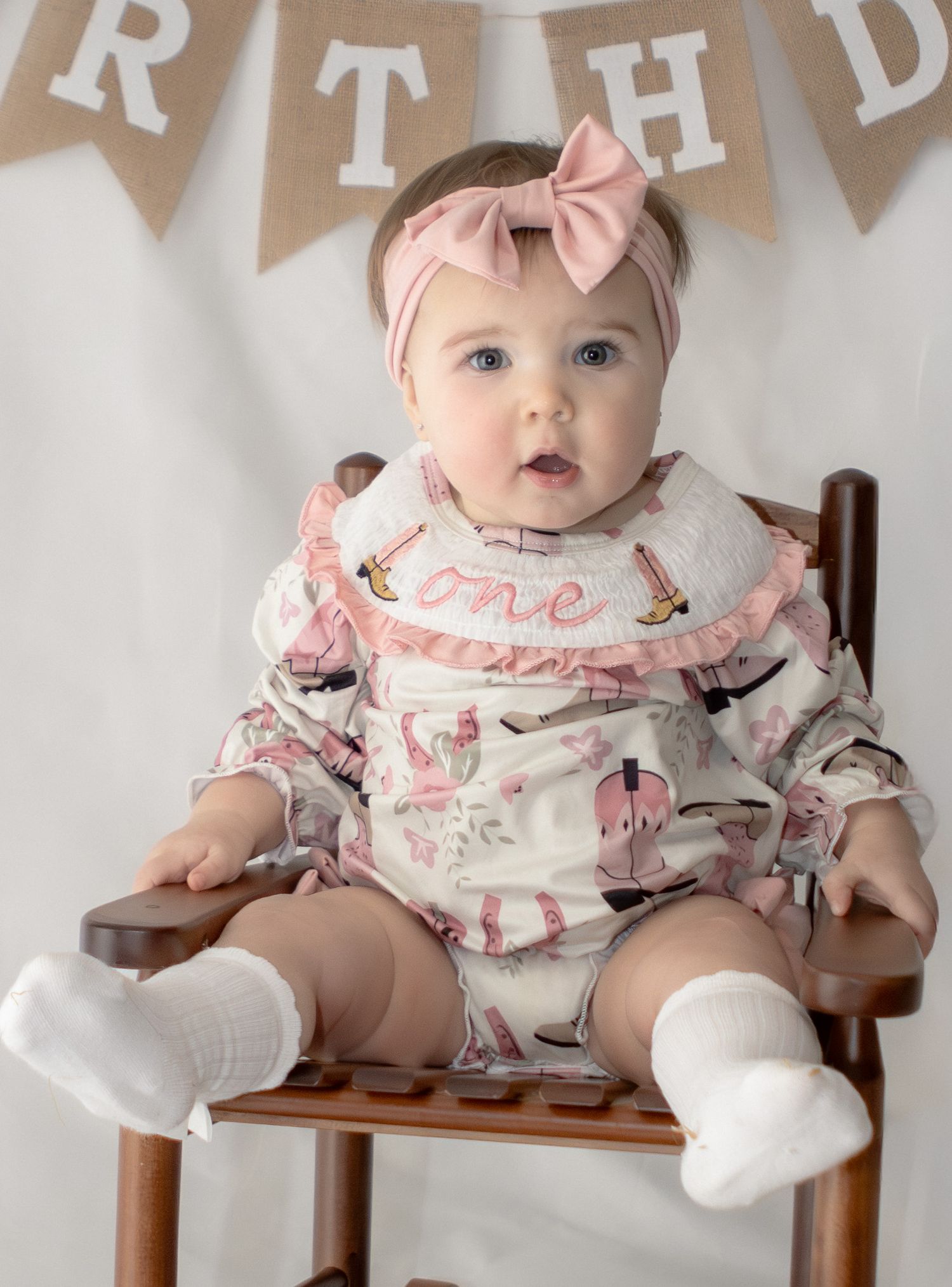 Baby girl in pink floral outfit with bow headband sitting in chair at first birthday party.