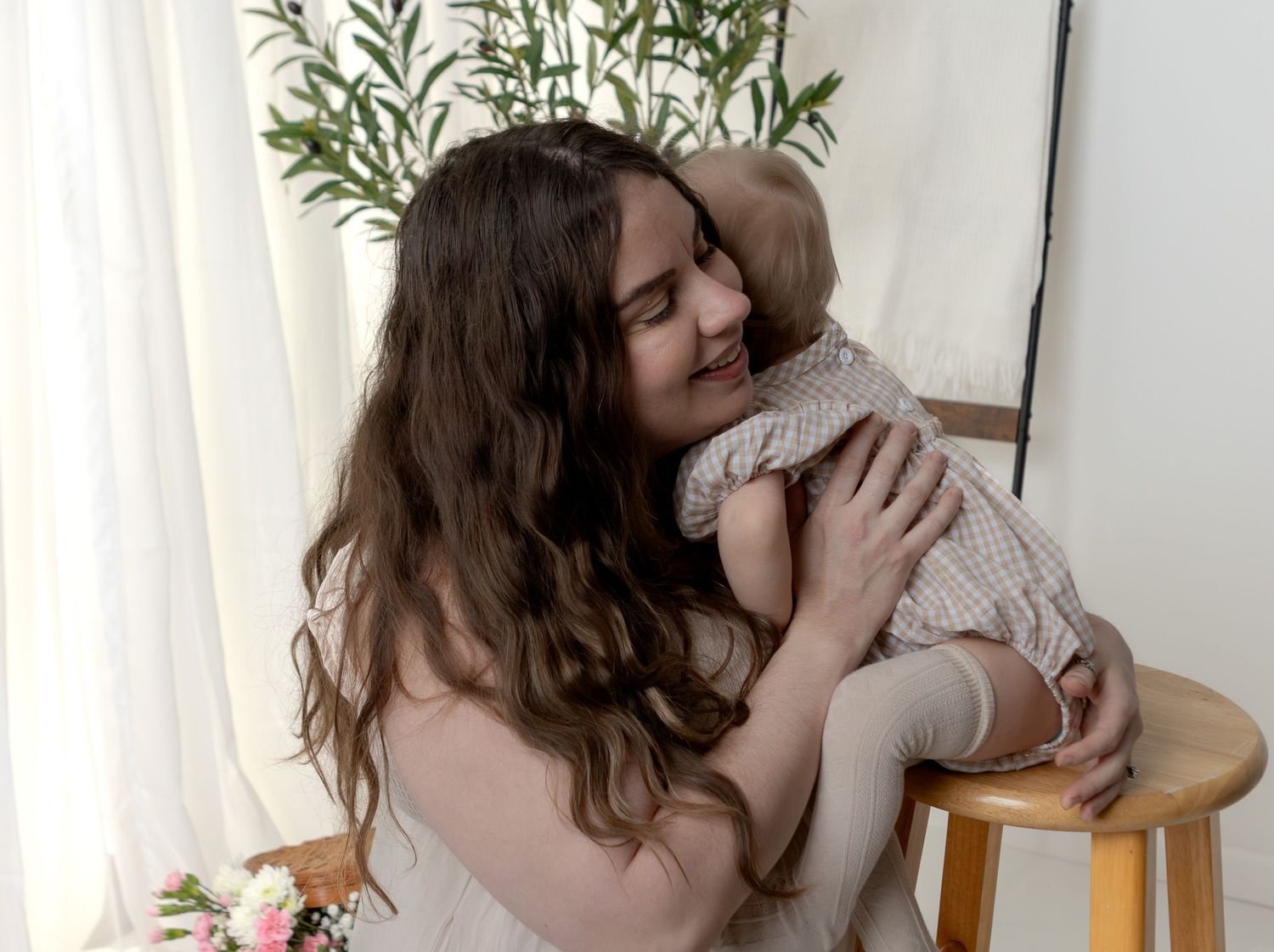 Young woman with long brown hair tenderly holds and smiles at a baby in a bright white studio setting.