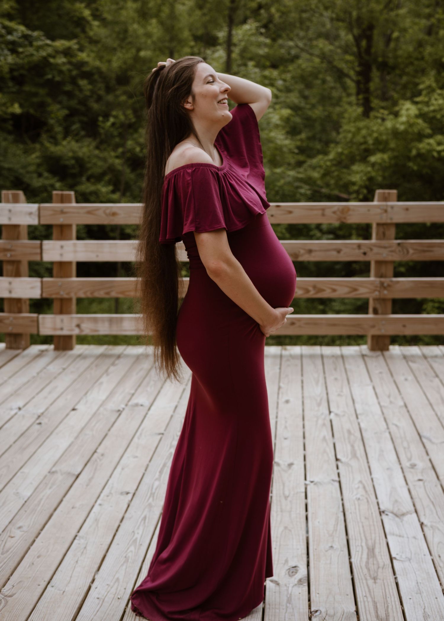 Pregnant woman in burgundy off-shoulder gown posing gracefully on an outdoor wooden deck.