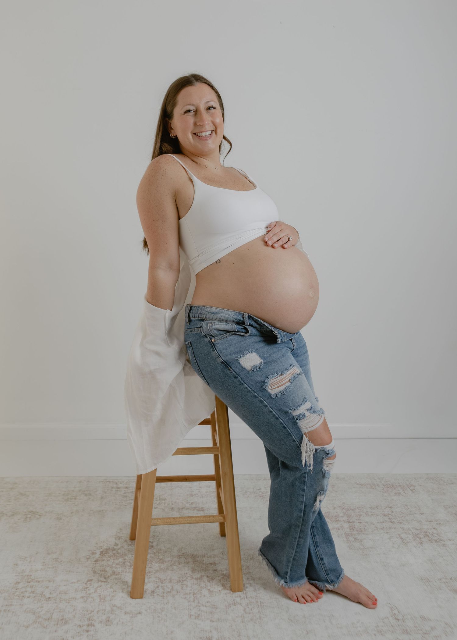 Smiling pregnant woman in white crop top and ripped jeans posing on wooden stool for maternity photo.