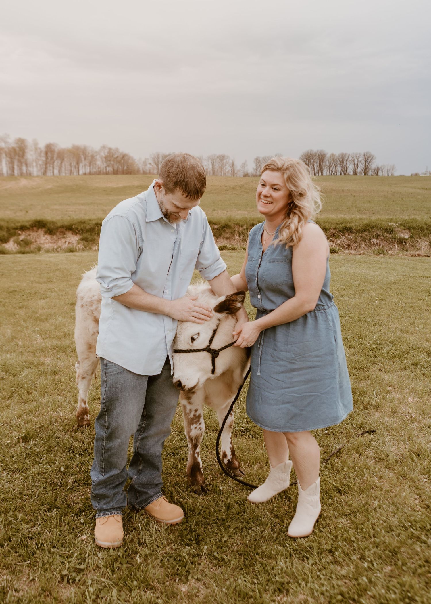 Couple petting a small goat in a green field during an outdoor farm engagement photo session.