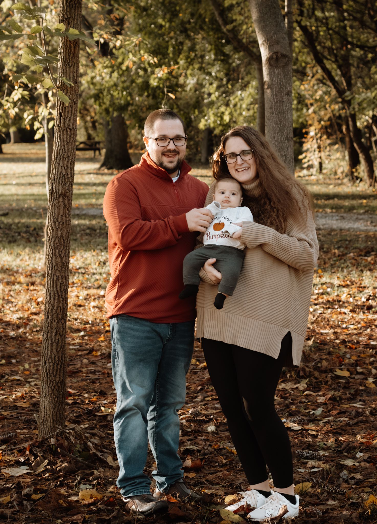 A couple stands among autumn trees holding their newborn baby dressed in a pumpkin onesie.