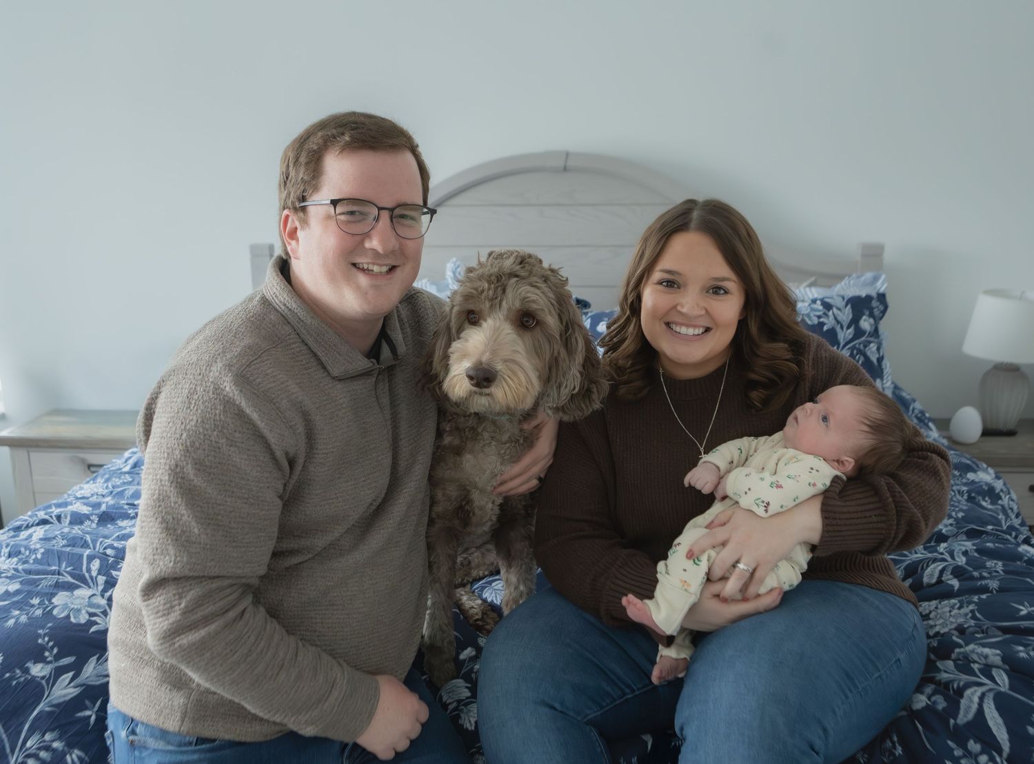 Happy family portrait with parents, curly-haired dog, and newborn baby sitting on a bed.