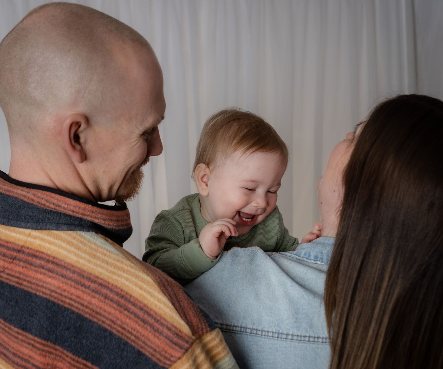 Laughing baby in green outfit is held by mother while father looks on warmly in soft light.