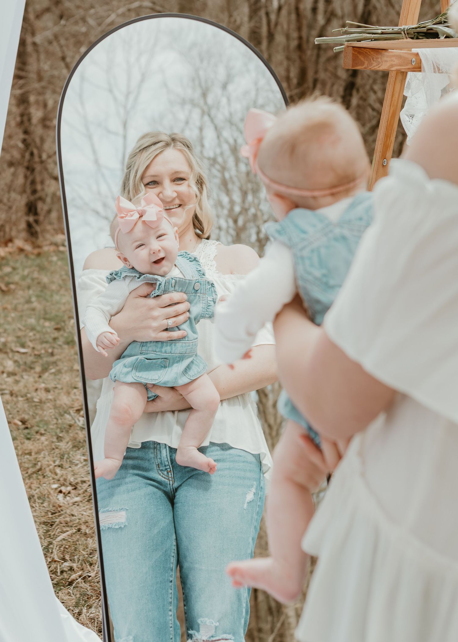 Blonde mother holding baby girl reflected in a tall arched mirror during outdoor session.