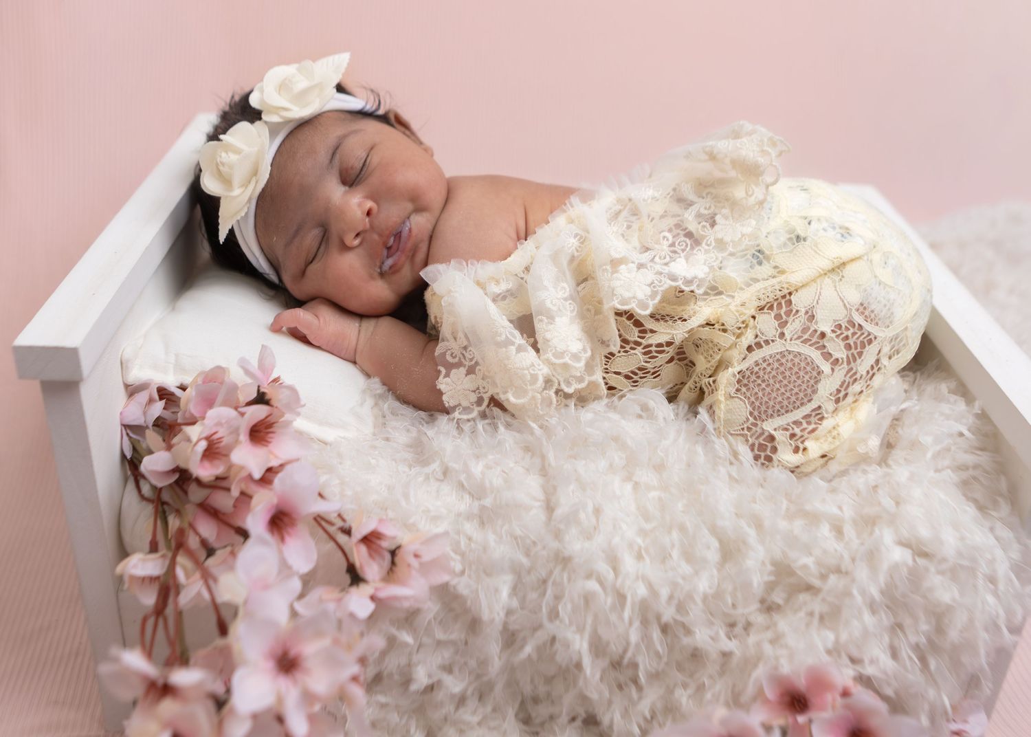Sleeping newborn in cream lace wrap with floral headband on fluffy white fur in white bed prop.
